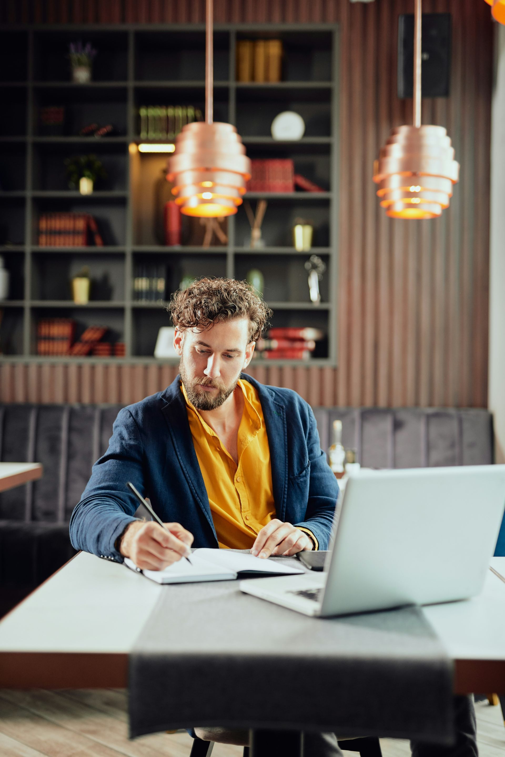 Man seated at a desk with a laptop and notebook in a modern office, writing under warm pendant lights