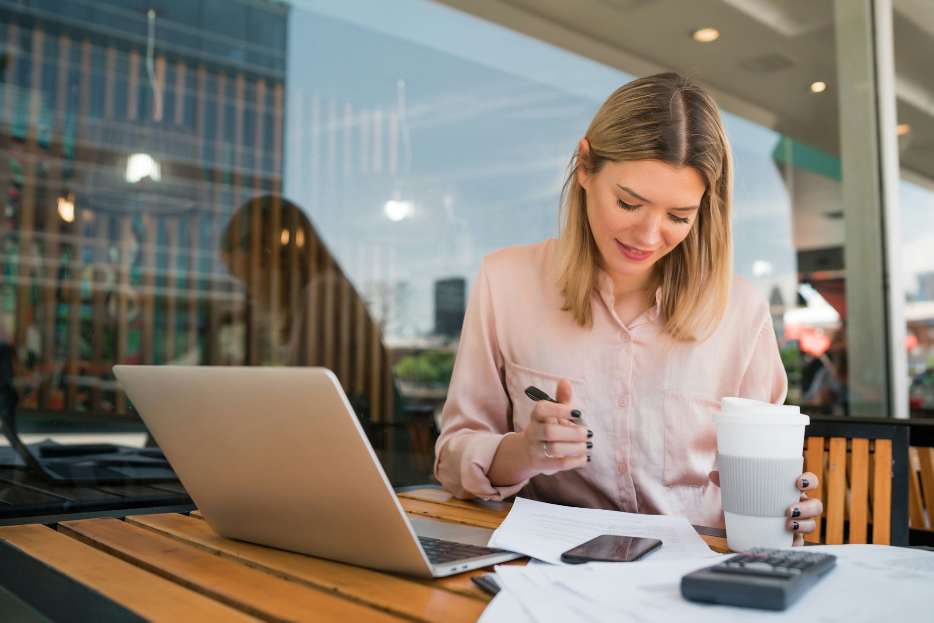 Woman working on a laptop at a café table with papers, a coffee cup, and a phone.