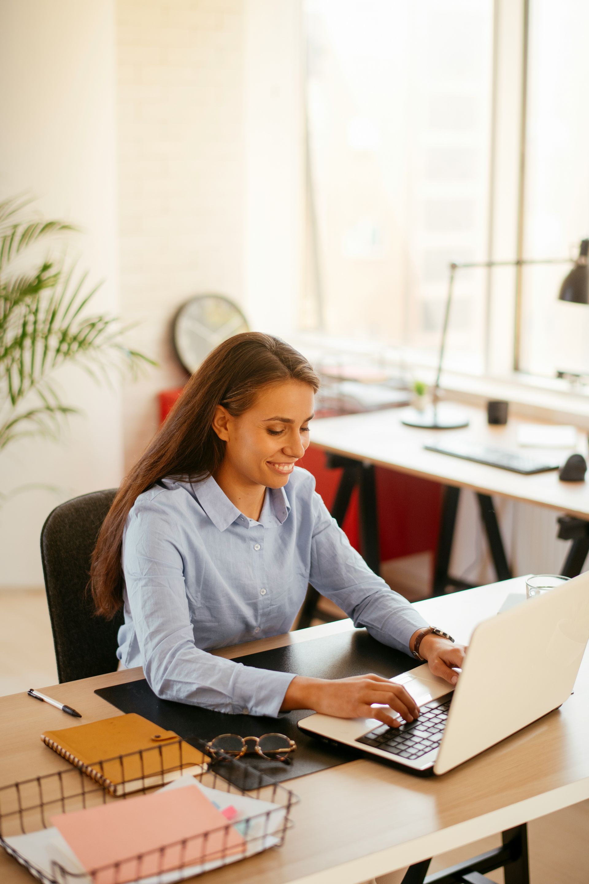 Woman working on a laptop at a desk in a bright office, taking notes beside a notebook and coffee mug