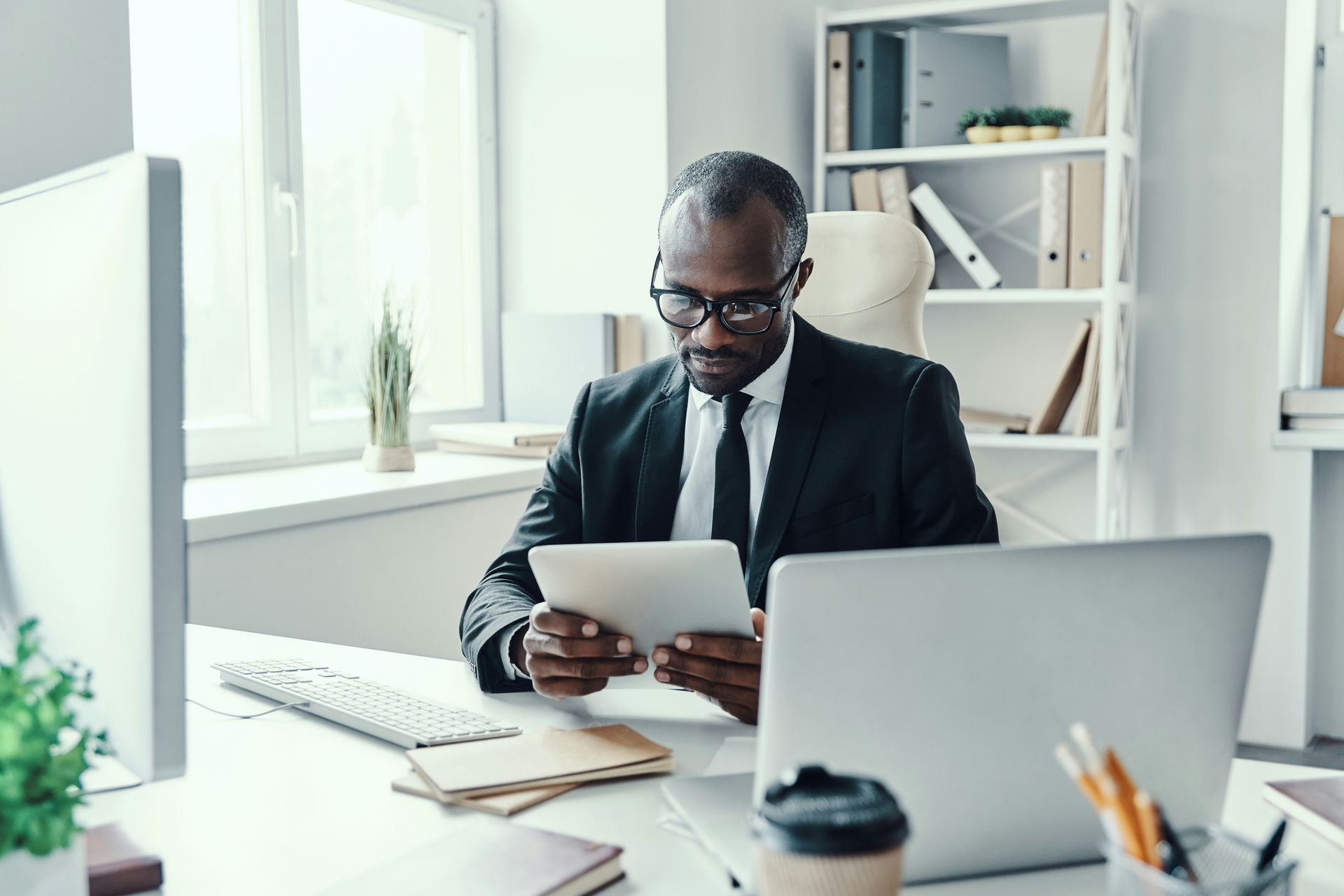 Person in a black suit using a tablet at a bright office desk with a laptop and coffee cup