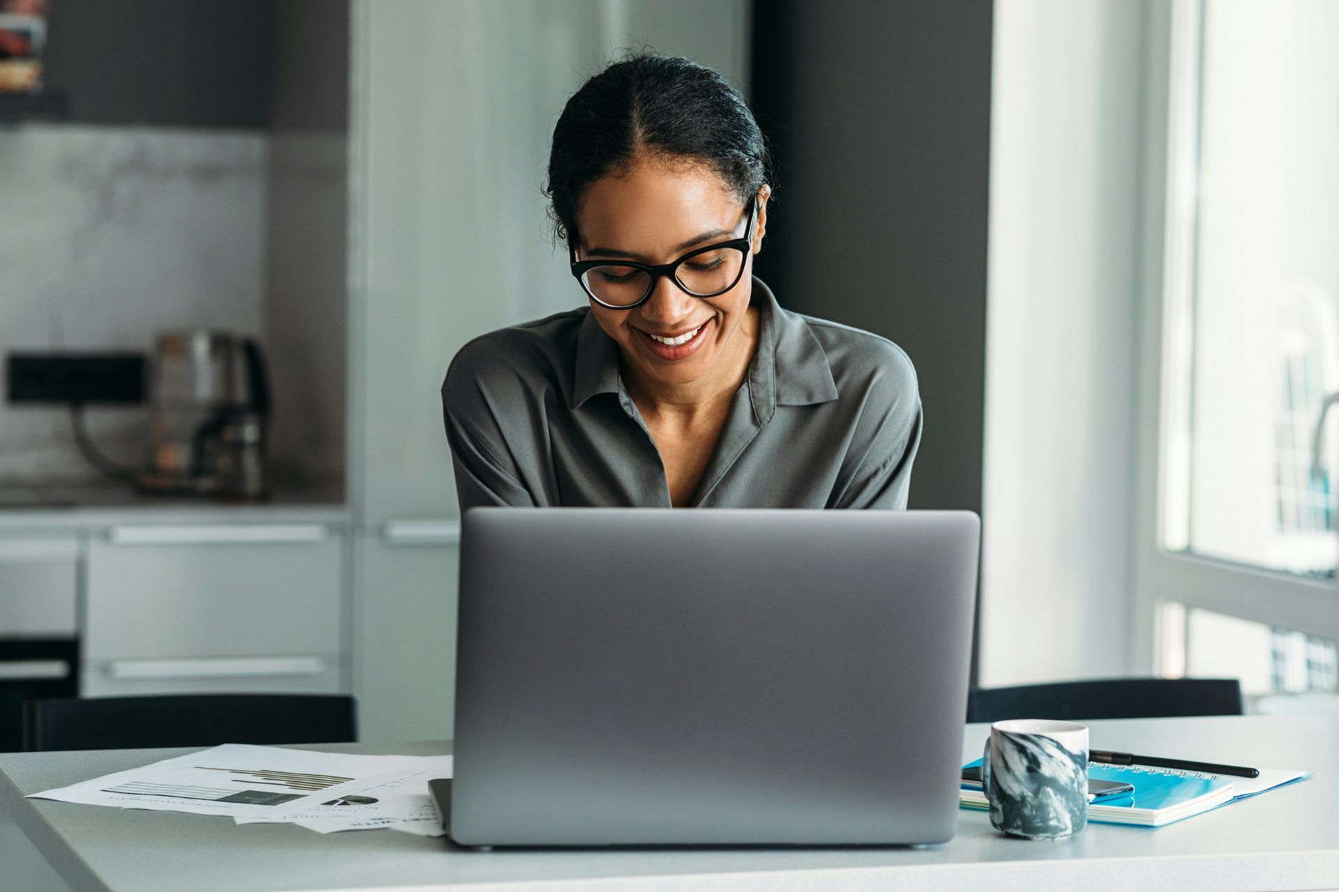 Smiling person working on a laptop at a kitchen table beside a window