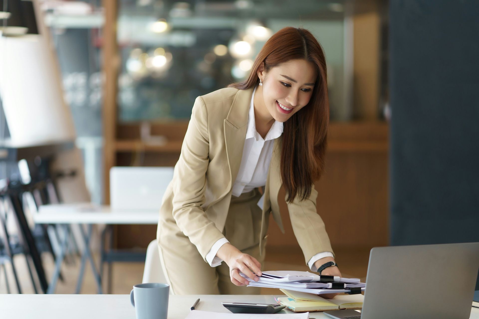 Woman in beige blazer leaning over a desk, reviewing papers beside a laptop in a modern office