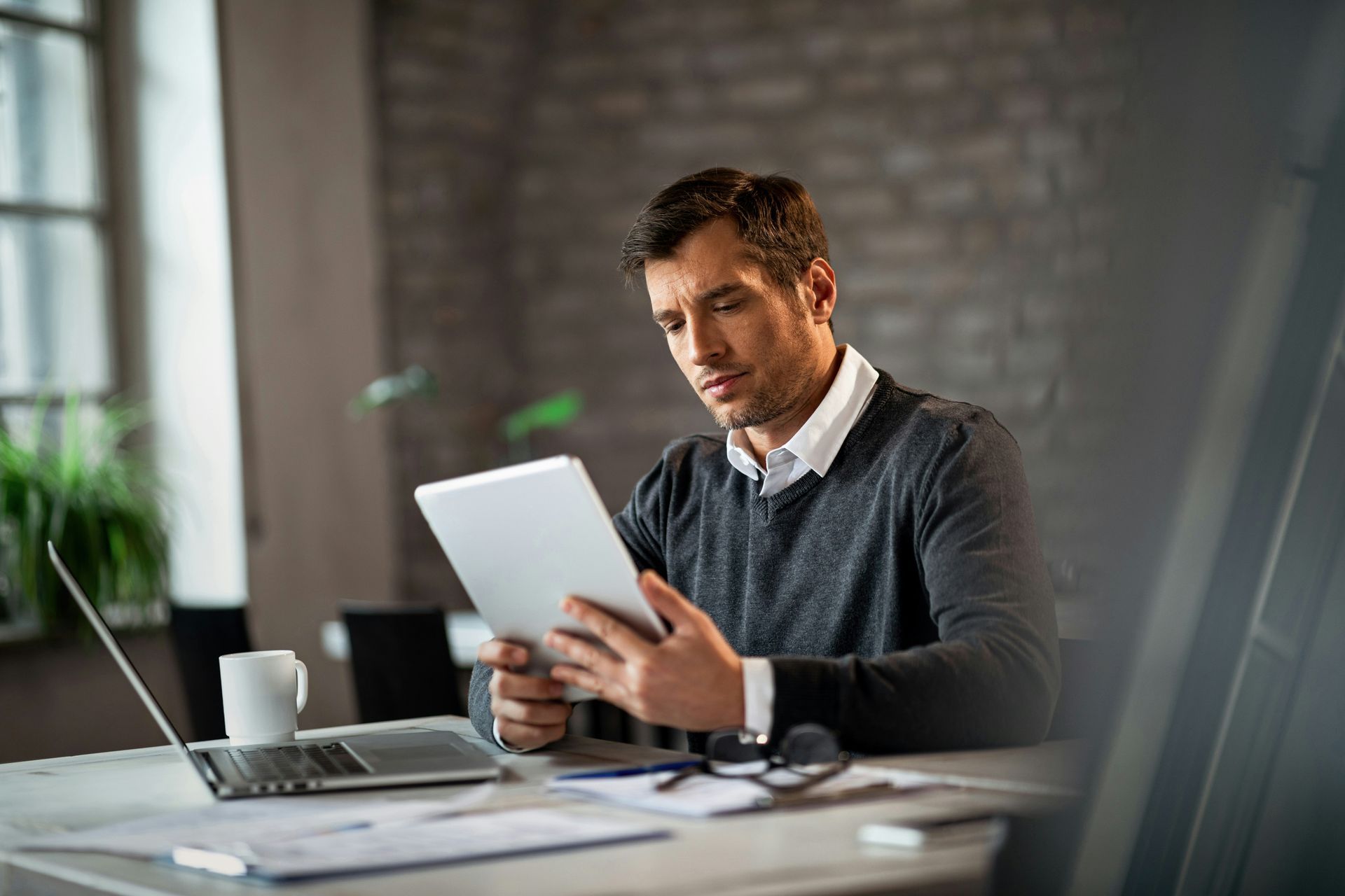 Man reviewing a tablet at a desk in a modern office, with a laptop and coffee cup nearby
