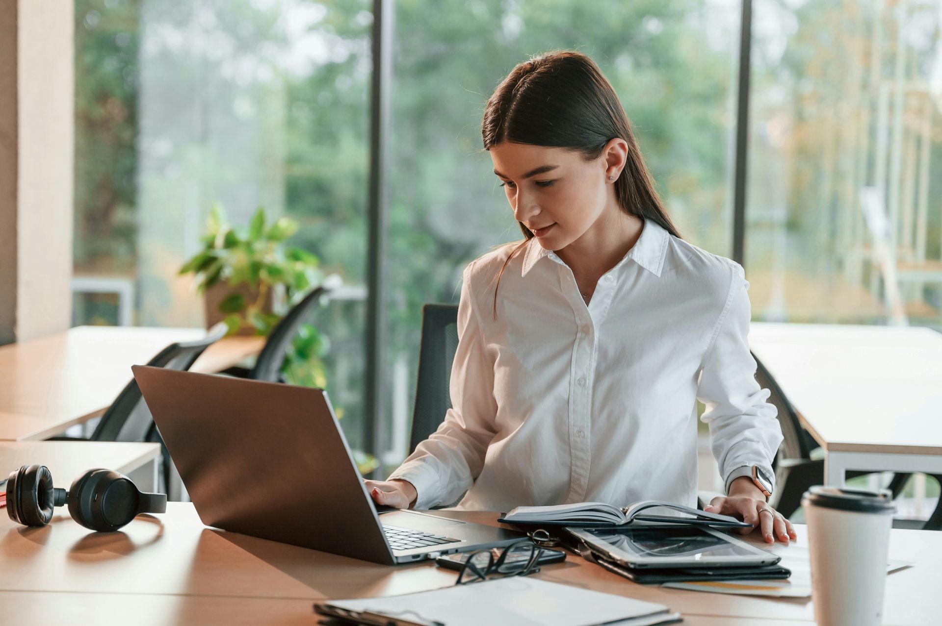 Woman working on a laptop at a desk with headphones, notebook, and coffee in a bright office