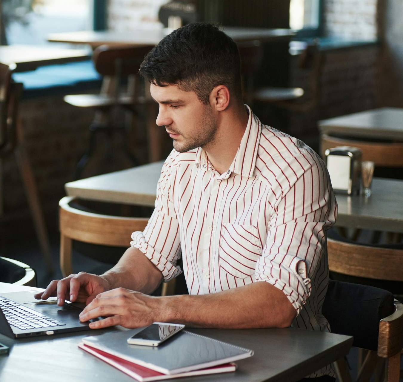Man typing on a laptop at a café table with a notebook and smartphone nearby
