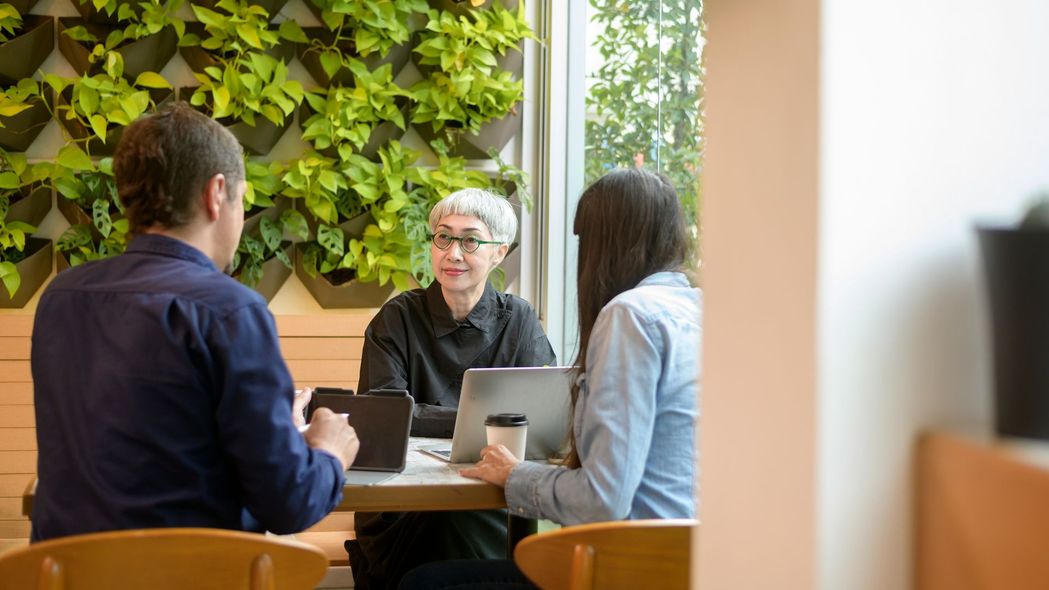 Three people in a cafe meeting with laptops and a coffee cup beside a green wall.