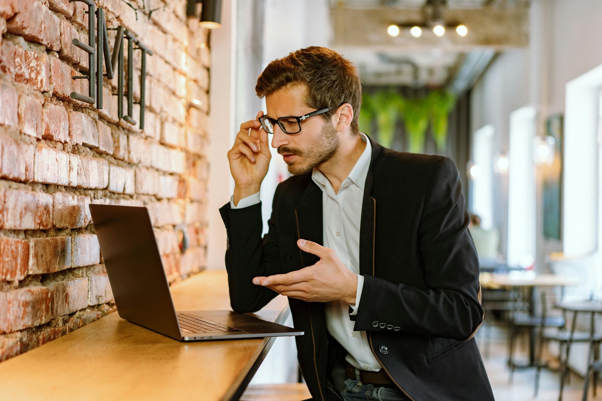 Man in a black suit adjusting glasses while using a laptop at a café counter