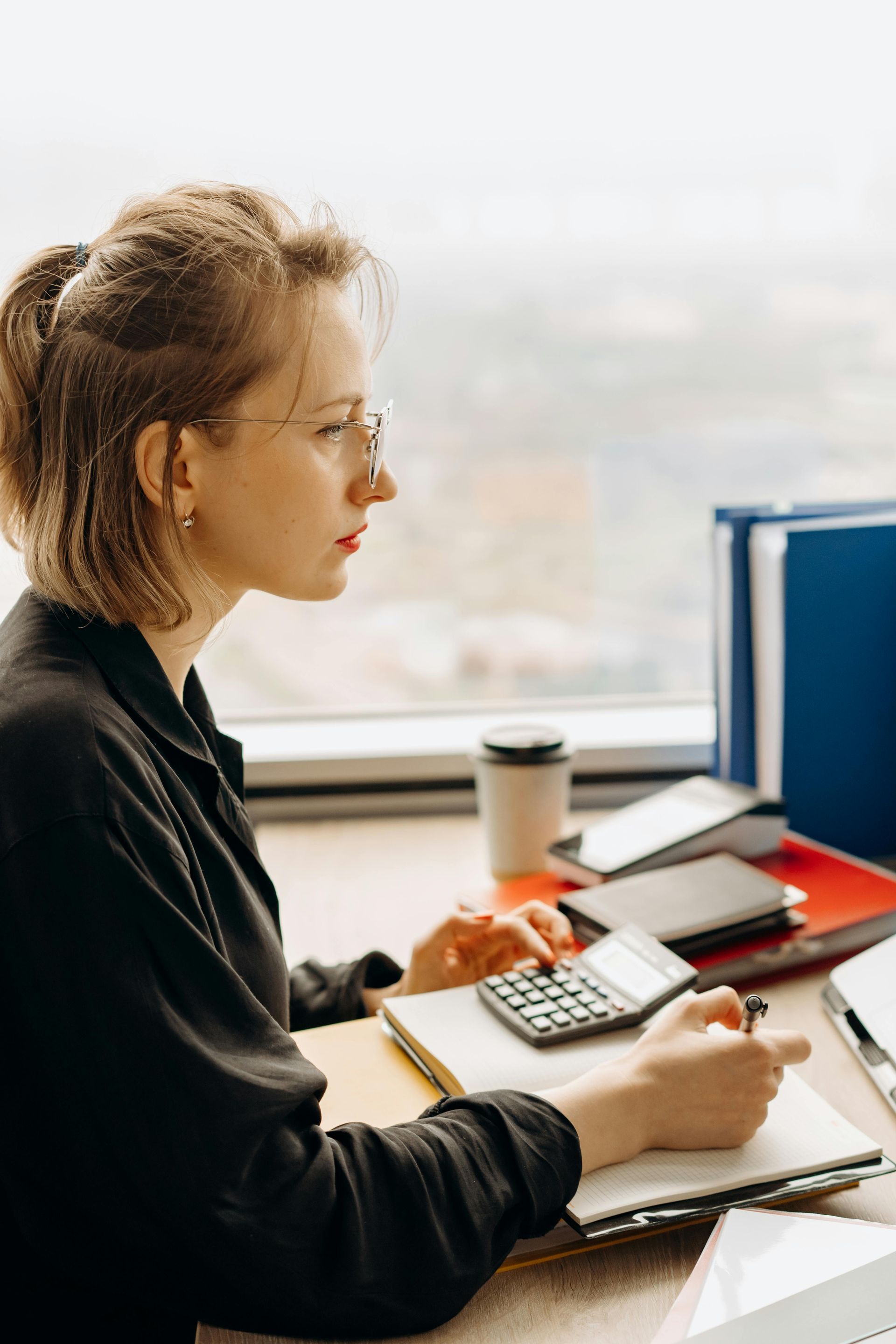 Person at a desk using a calculator and writing notes beside a window.