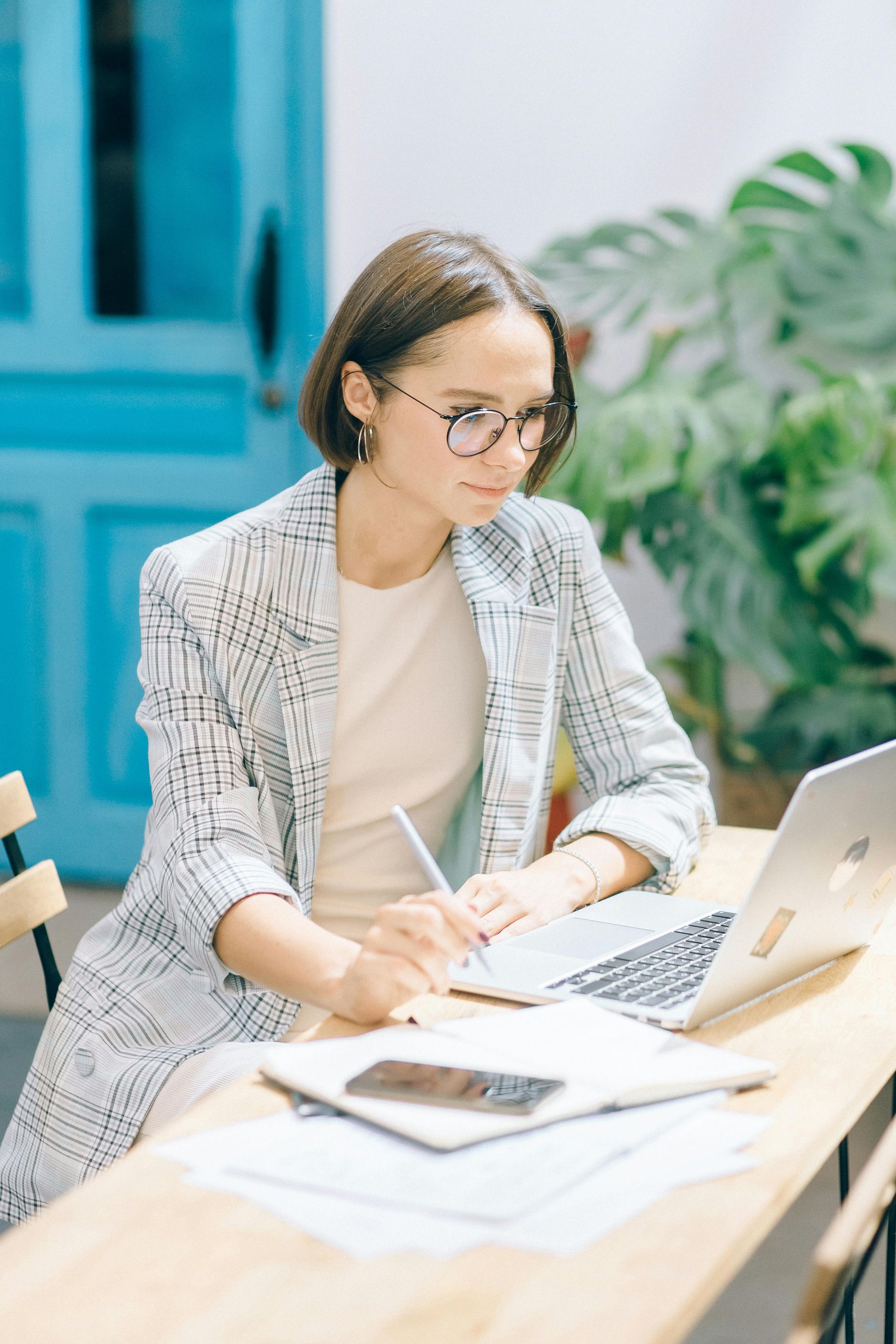 Woman working at a laptop in a bright office, writing notes at a desk.