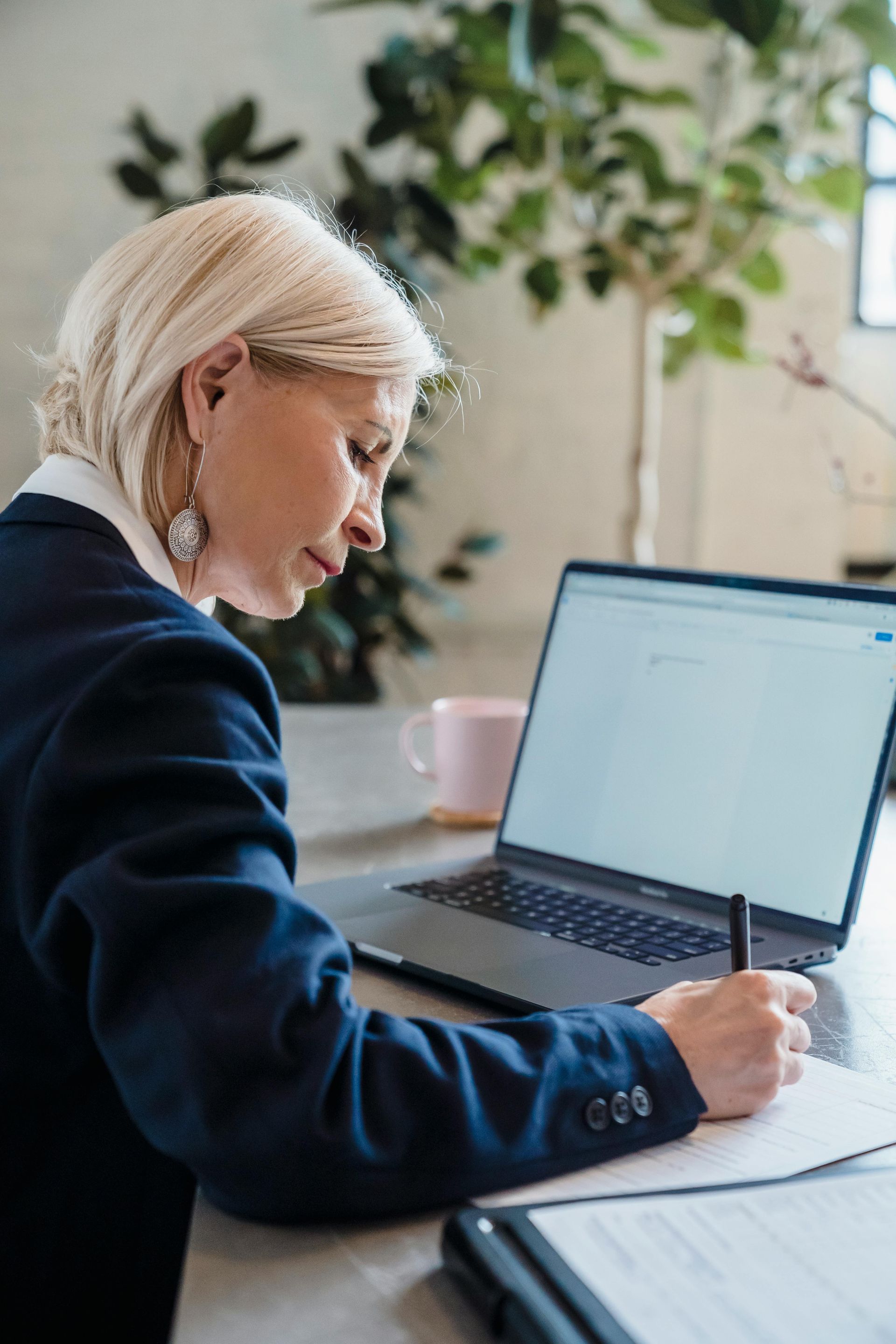 Person taking notes beside an open laptop at a desk in a bright office