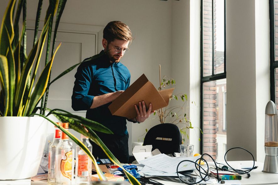Man in blue shirt unpacking a cardboard box beside a bright window, with plants and desk cables nearby