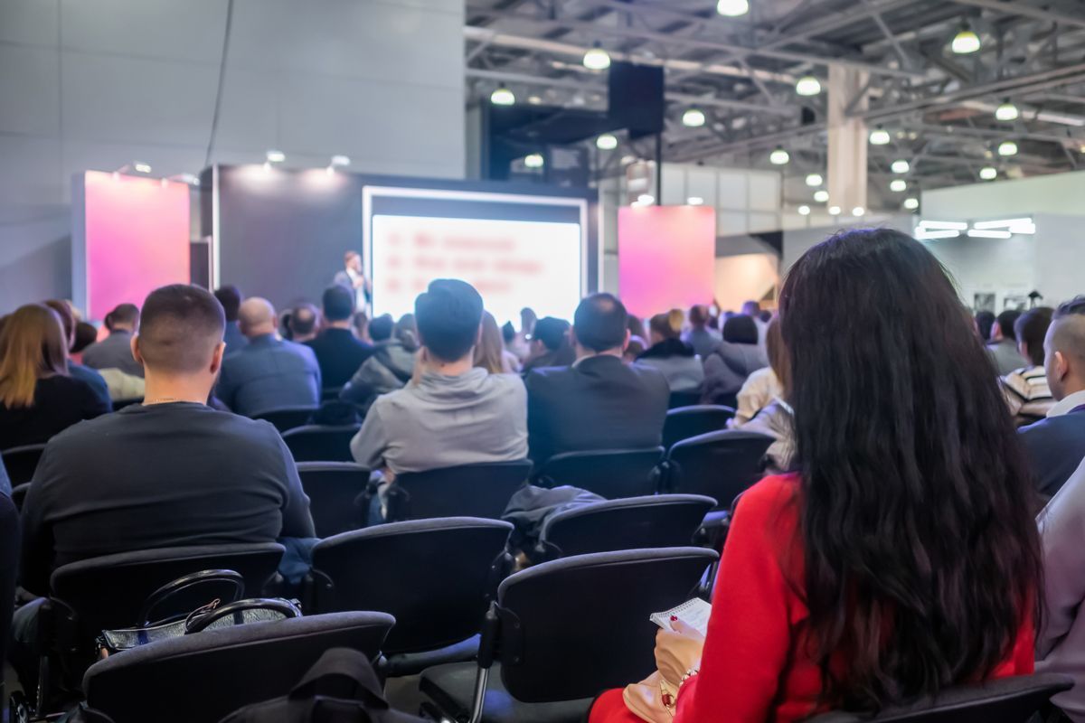 Audience seated at an indoor conference, facing a bright presentation screen.