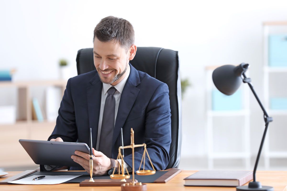 Man in a suit using a tablet at a desk with a balance scale and lamp in a bright office