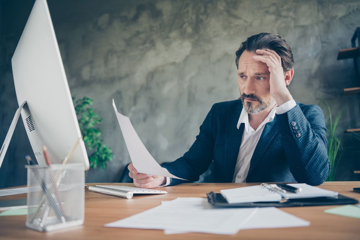 Man in a dark blazer reviews papers at a desk with a laptop and monitor in a bright office.