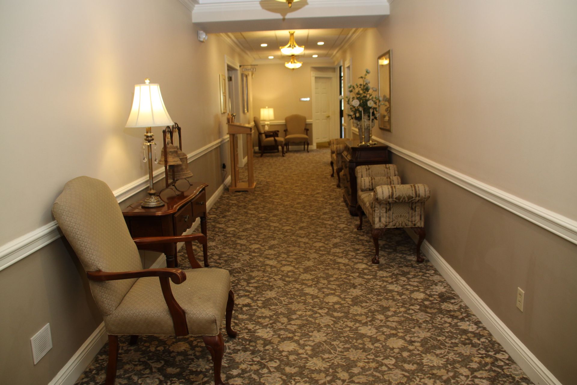 Empty hotel hallway with patterned carpet, beige walls, chairs, and a table lamp