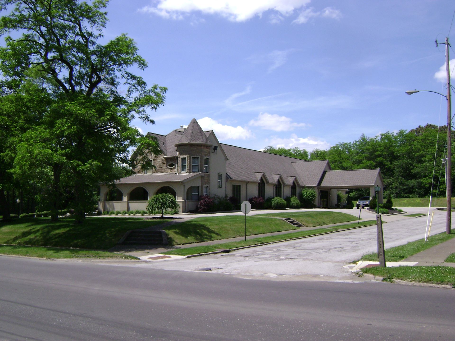 Large stone building with arched porch on a tree-lined street corner under a blue sky