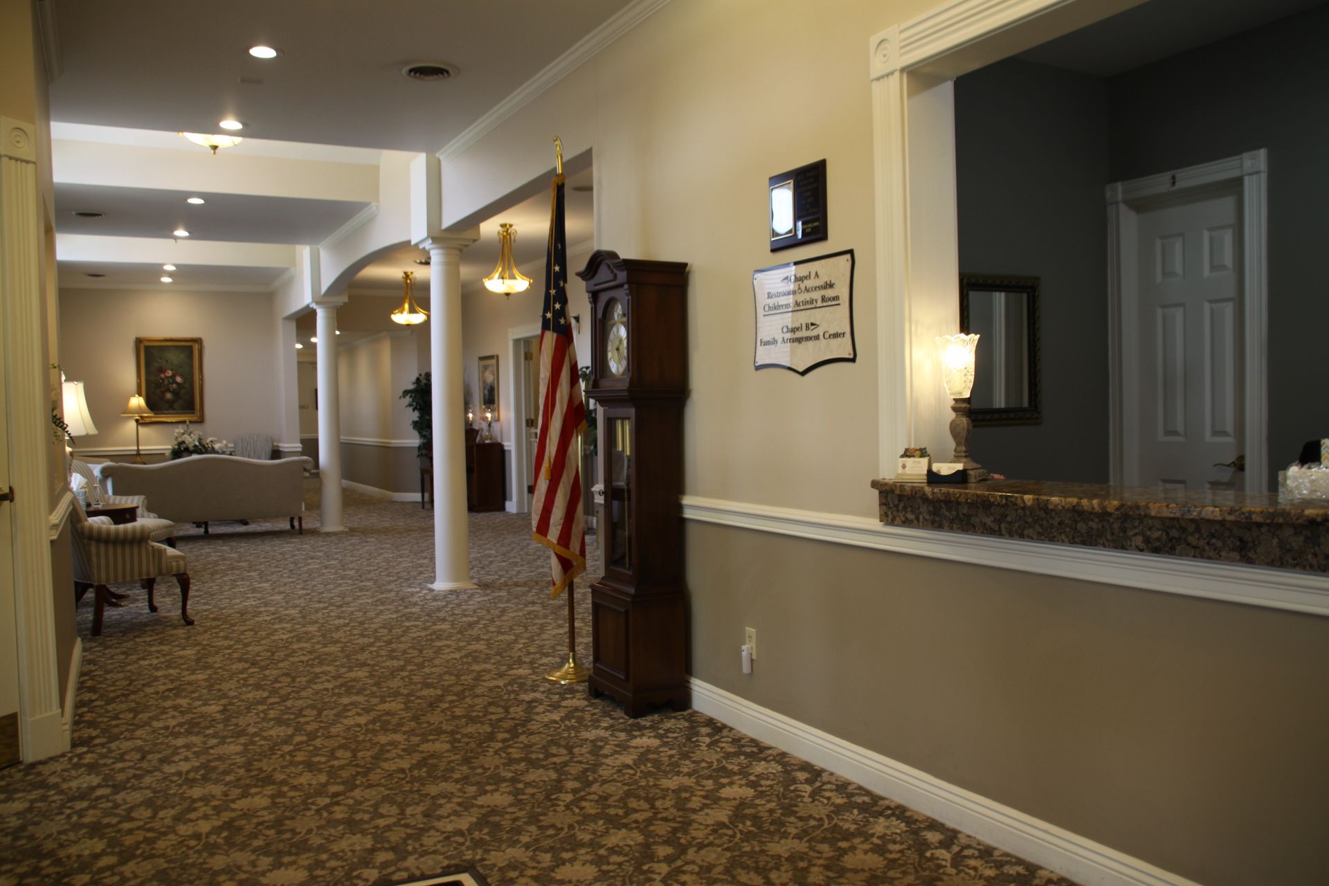 Hotel hallway with patterned carpet, wall sconces, and a reception window counter on the right.