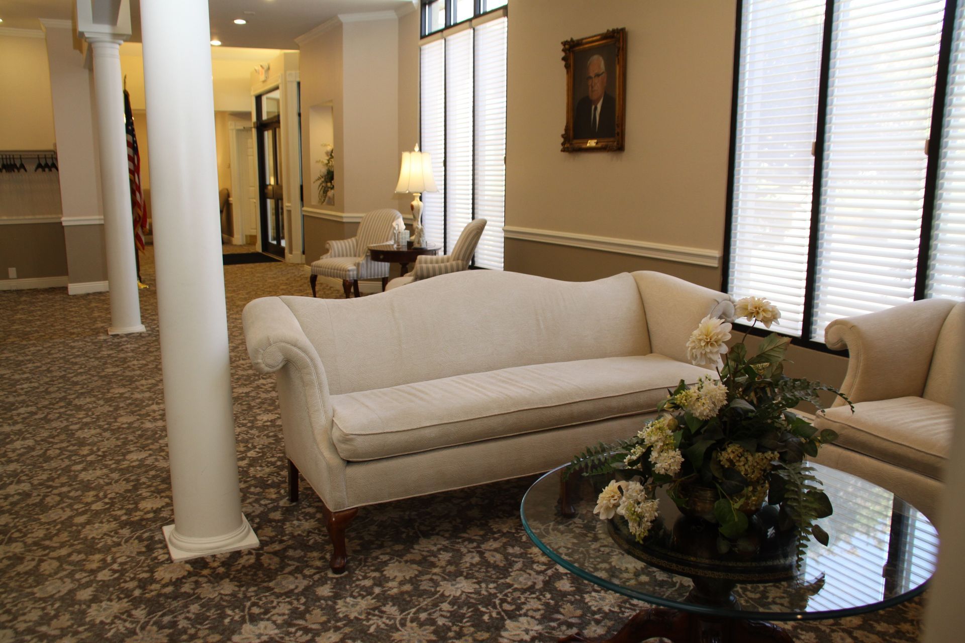Elegant living room with white sofas, glass coffee table, floral arrangement, and tall windows with blinds