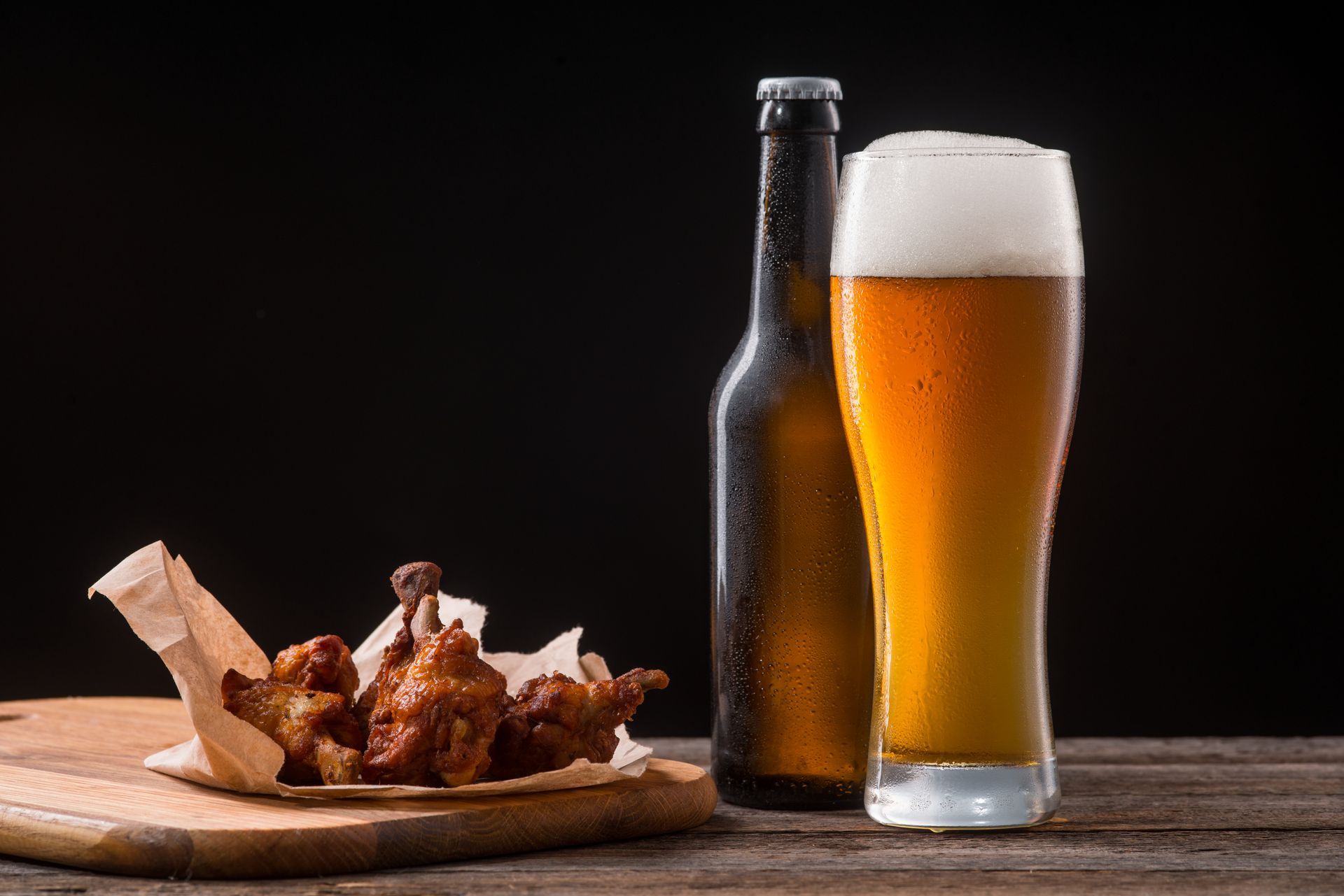 Chicken wings on a wooden board next to a beer bottle and glass of beer, on a wooden table.