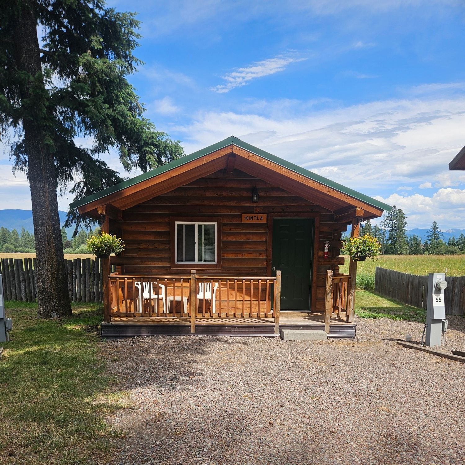 A small wooden cabin with a porch, green door, and window under a blue sky, surrounded by grass and gravel.