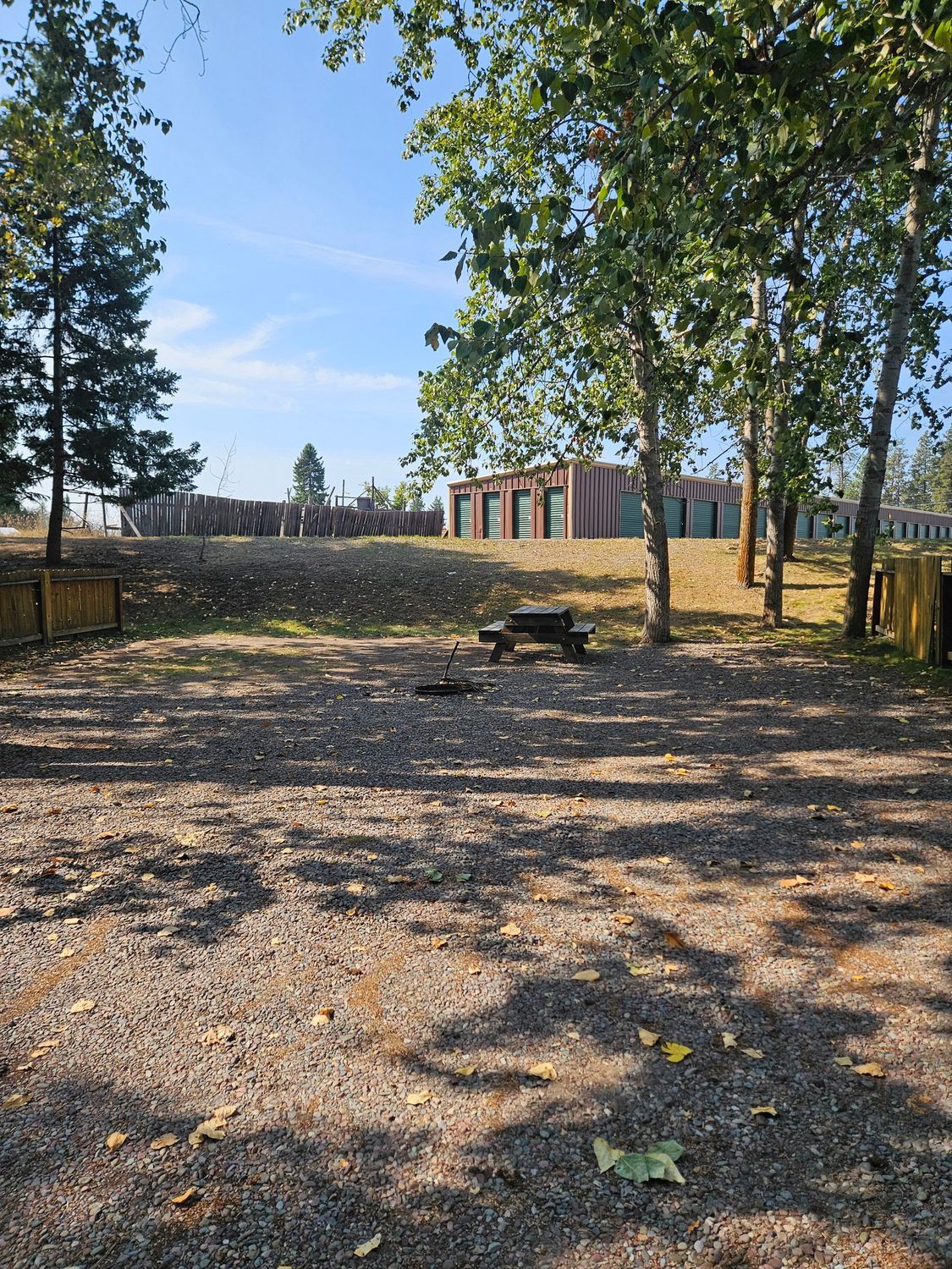 Gravel area with picnic table, fire pit, and trees; modern building in the background under a blue sky.