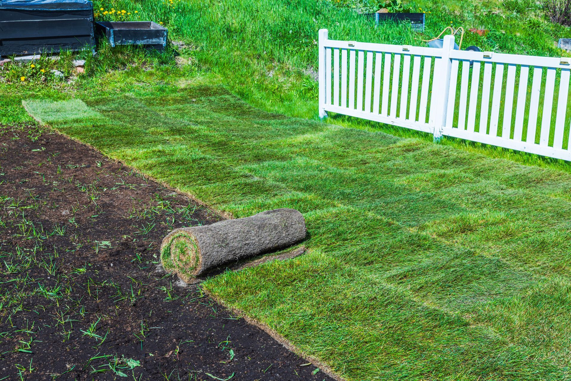 Freshly rolled sod beside a dark soil patch in a yard next to a white fence
