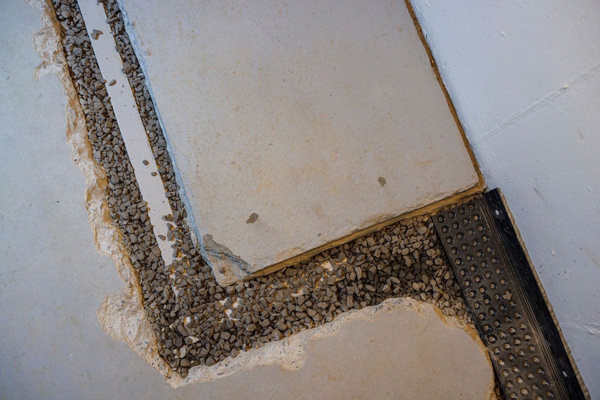 Concrete and gravel flooring under construction, with a partially removed tile showing the substrate and drainage system.