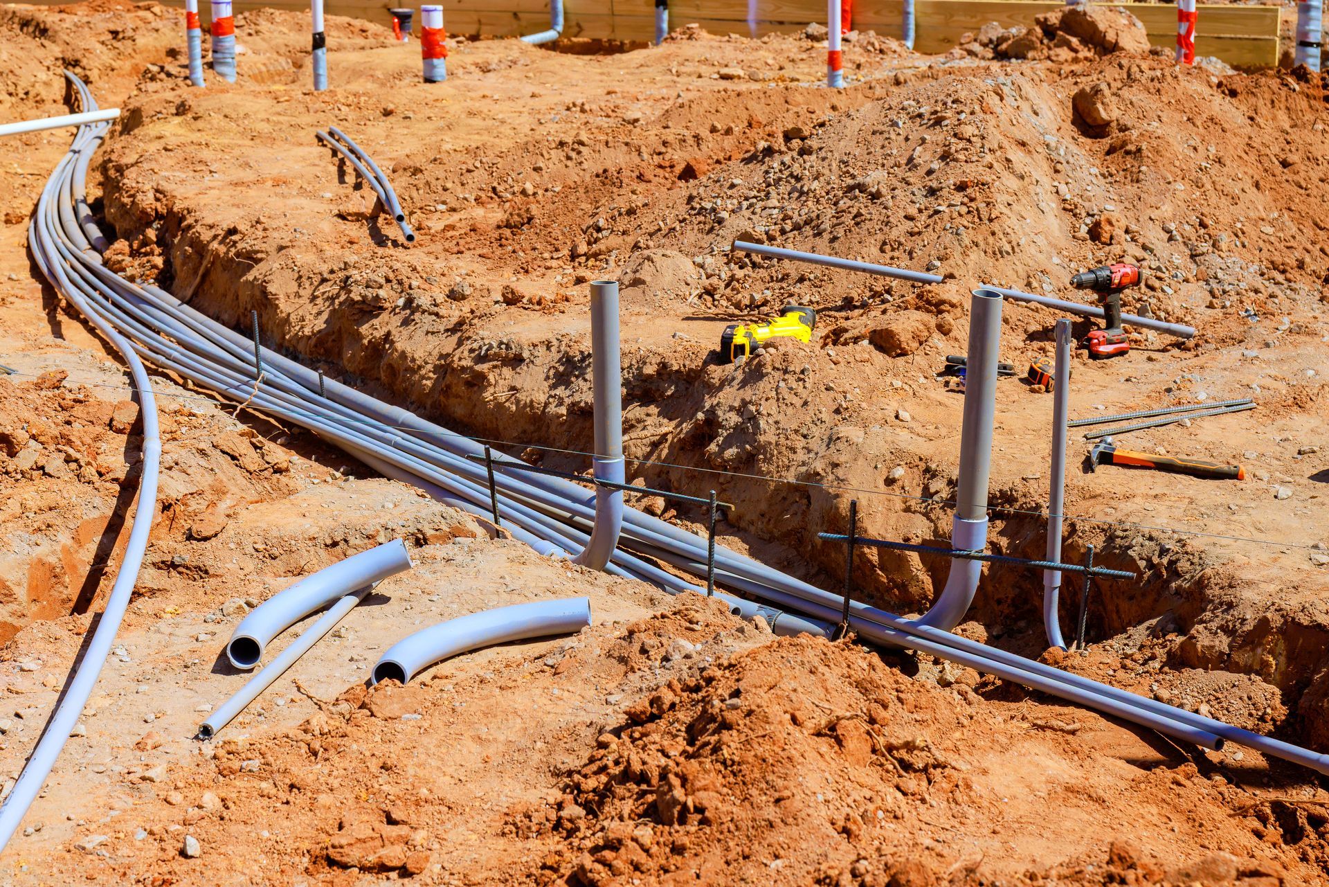 Trench with gray electrical conduits and PVC pipes in brown earth at a construction site.