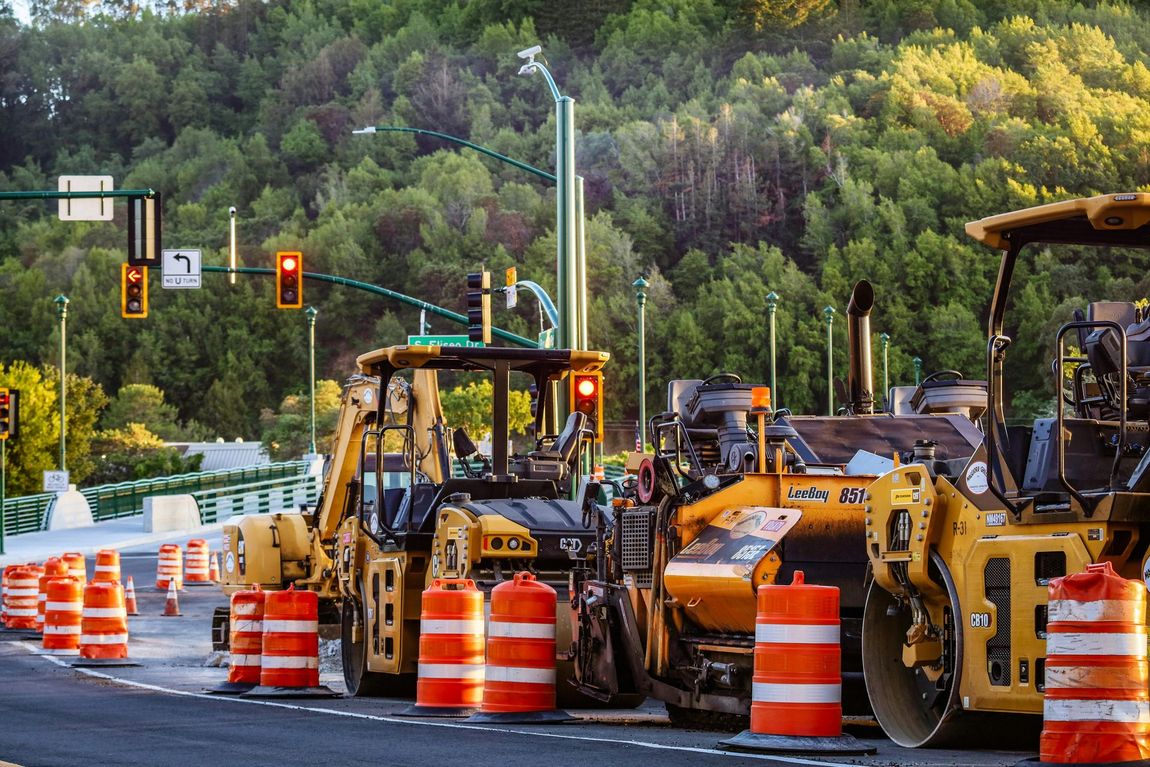 Construction site with yellow machinery and orange barrels; traffic lights with red signals; trees in background.