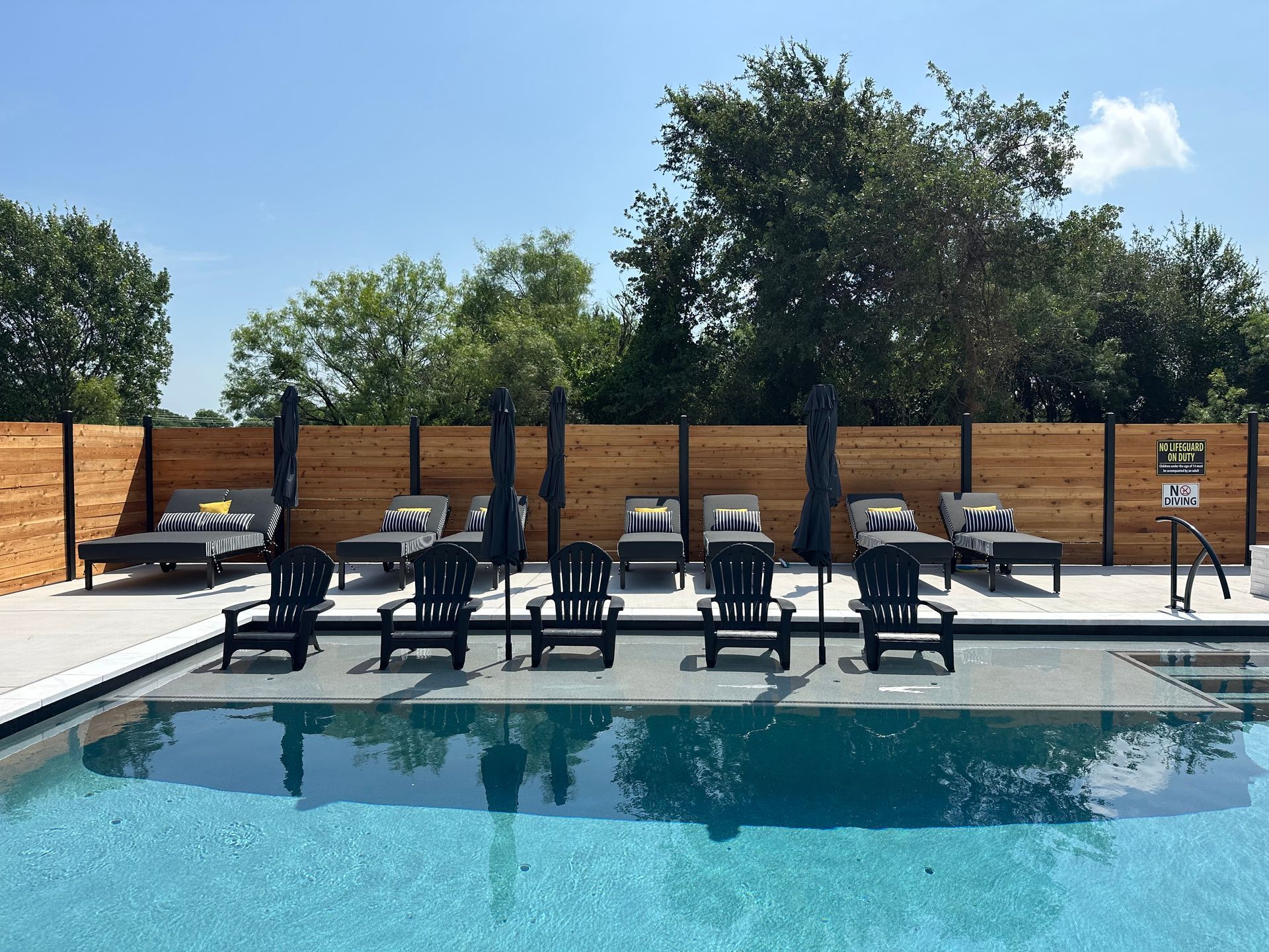 Backyard pool with black loungers, brick fence, and trees under a sunny sky