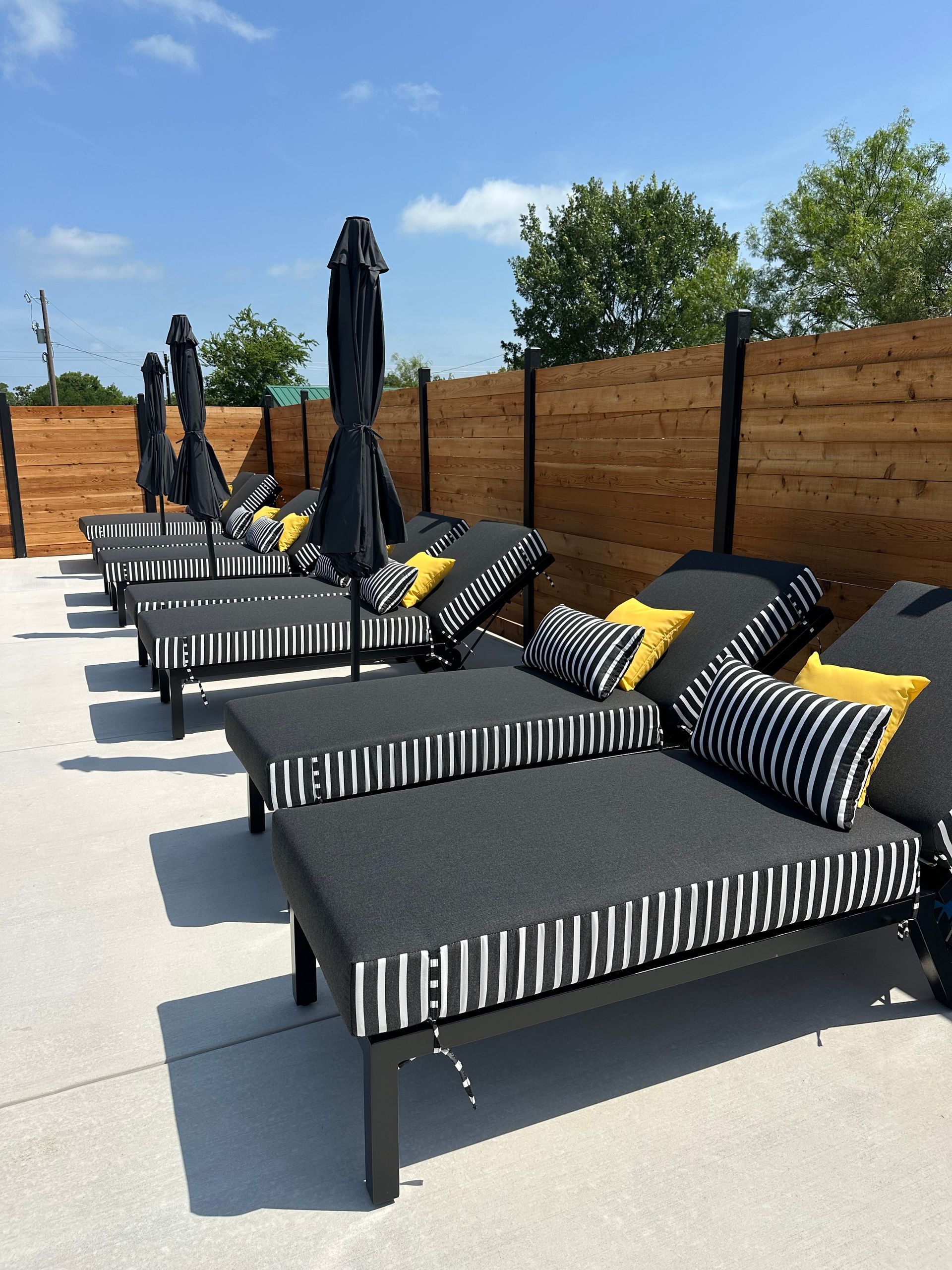 Row of black lounge chairs with striped cushions and yellow pillows on a sunny outdoor patio with closed umbrellas
