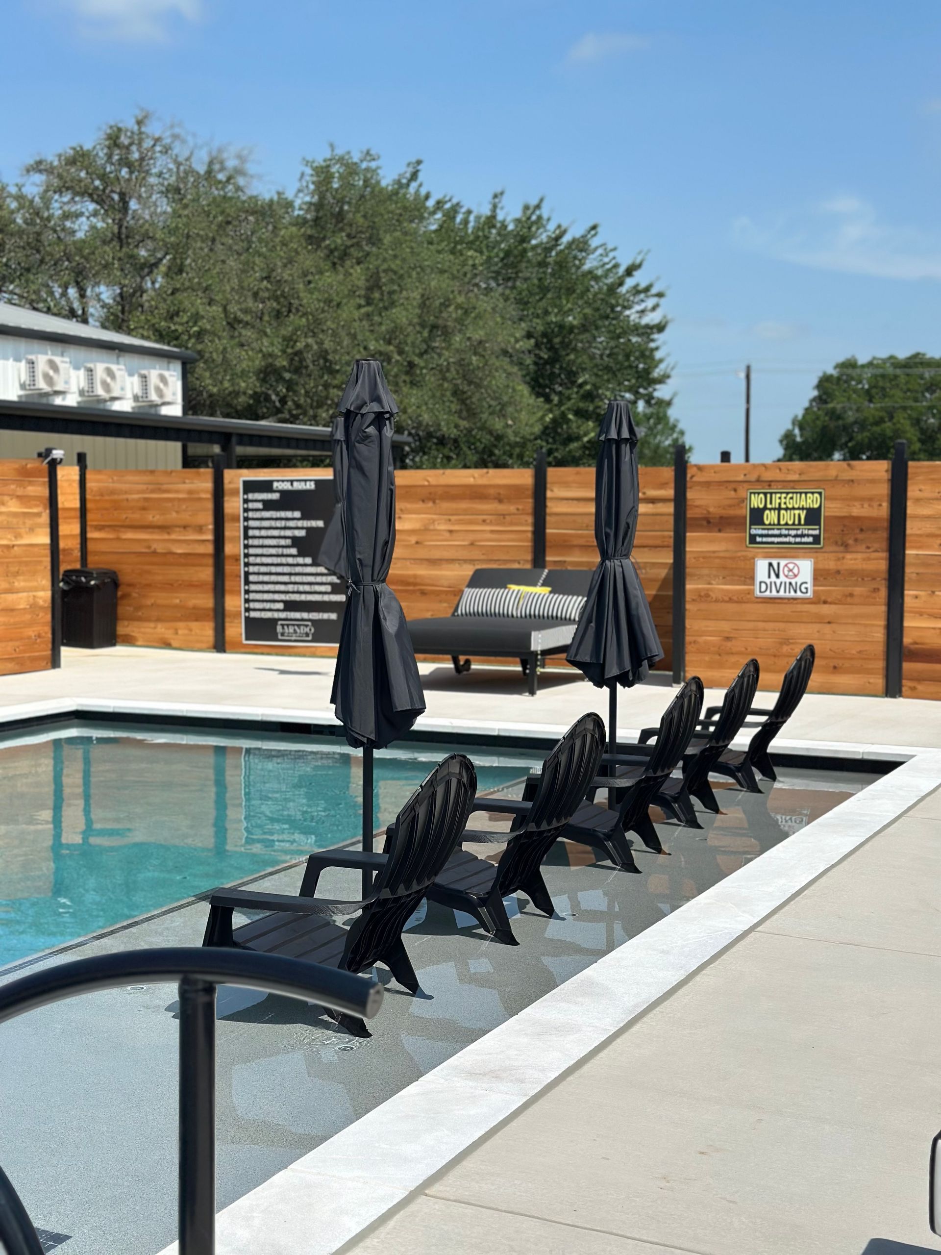 Outdoor pool with black lounge chairs, closed umbrellas, and a wooden privacy fence under a blue sky