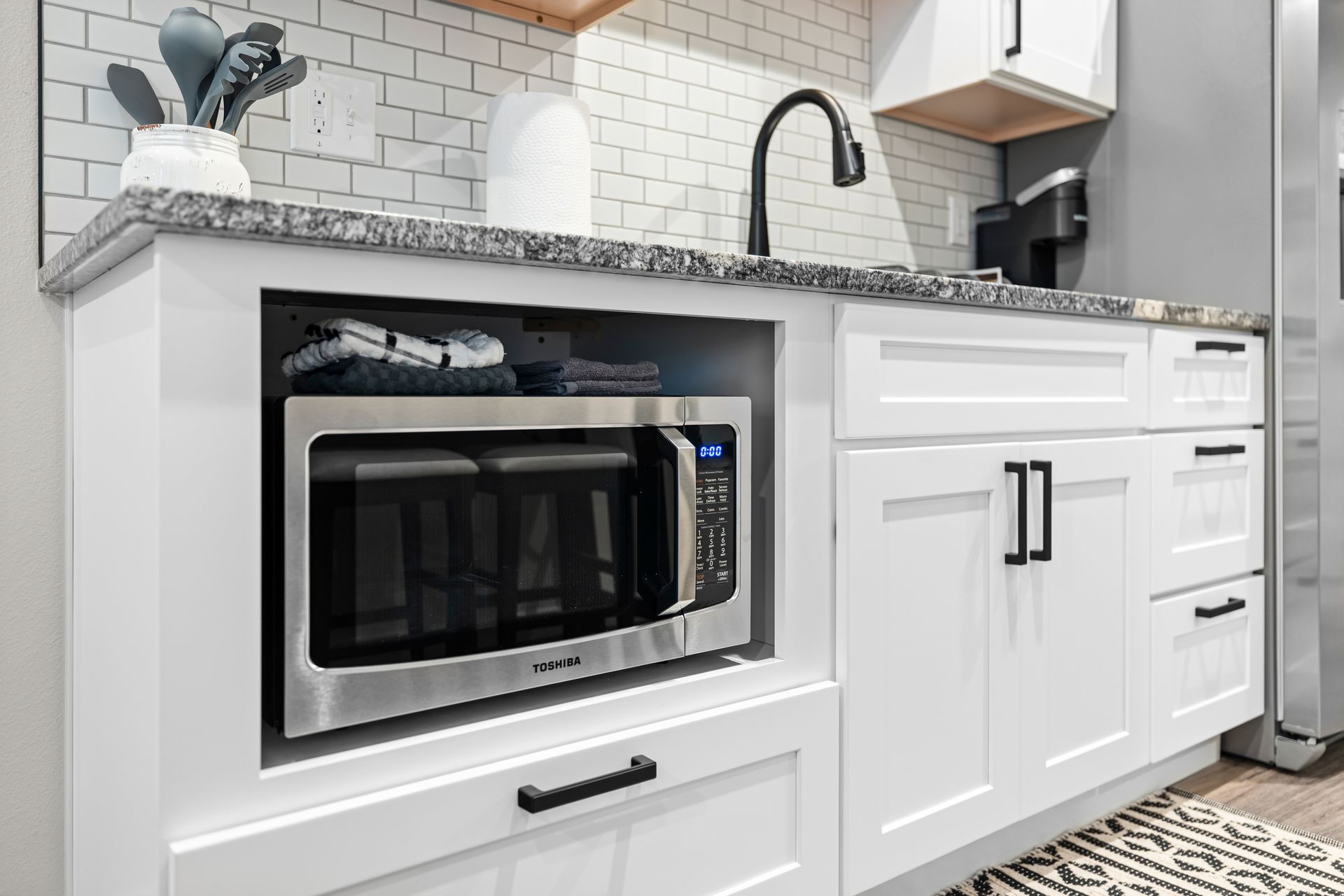 White kitchen with granite countertops, stainless microwave, sink, and white cabinets with black handles