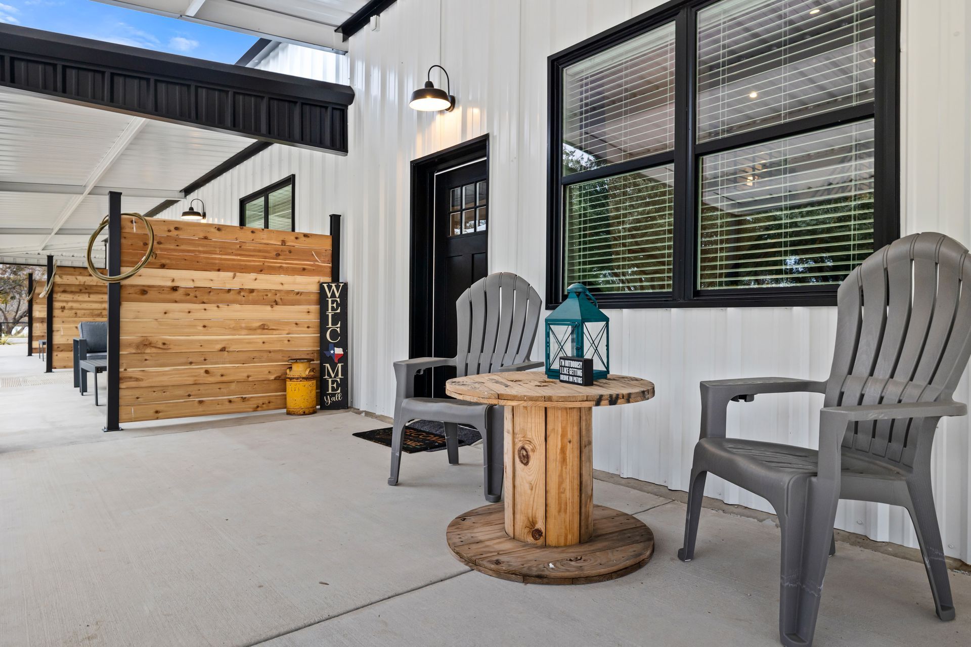Covered patio with two gray chairs and a round wooden table beside a wooden gate and black-framed window