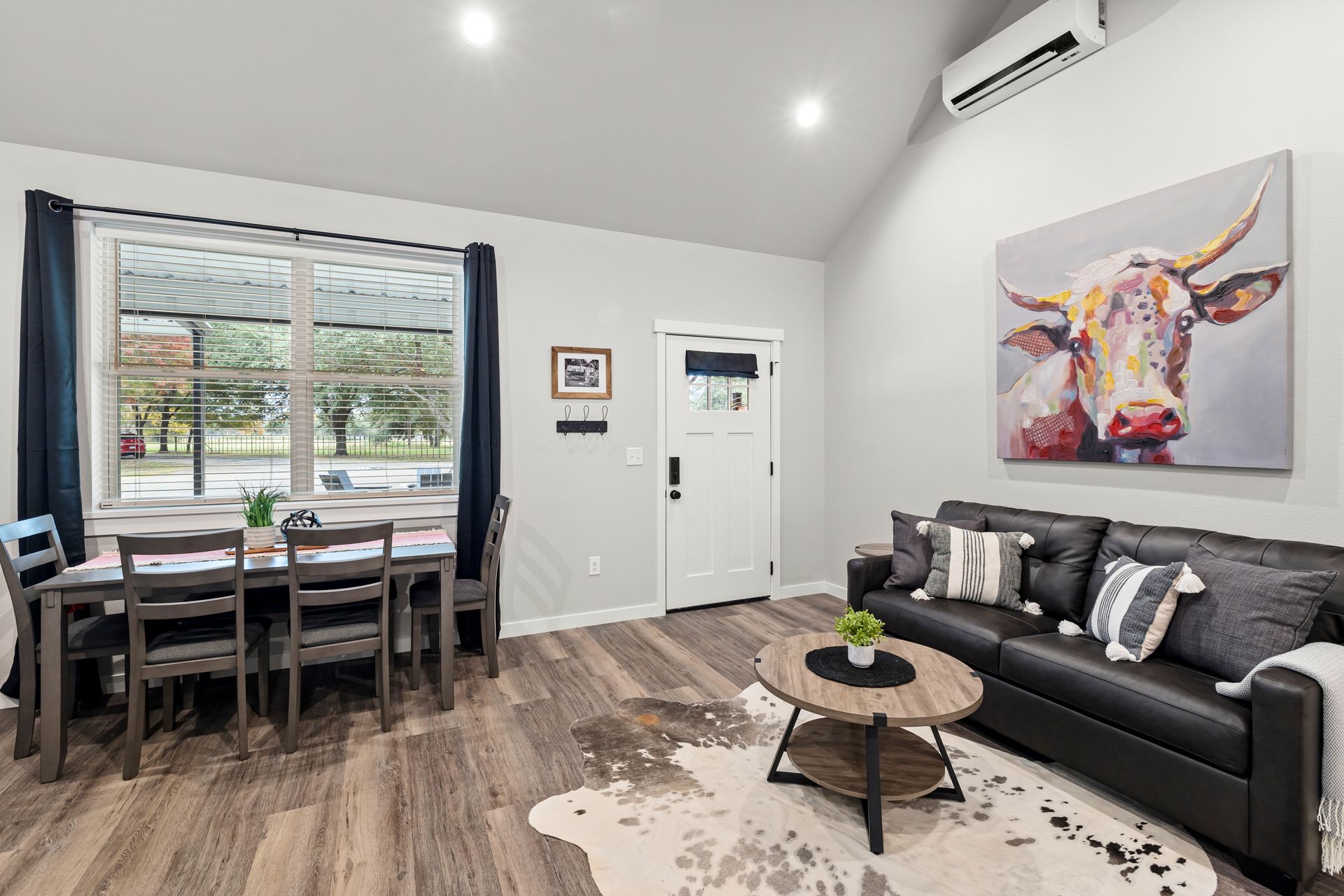 Bright living room with black sofa, dining table, large window, and colorful wall art