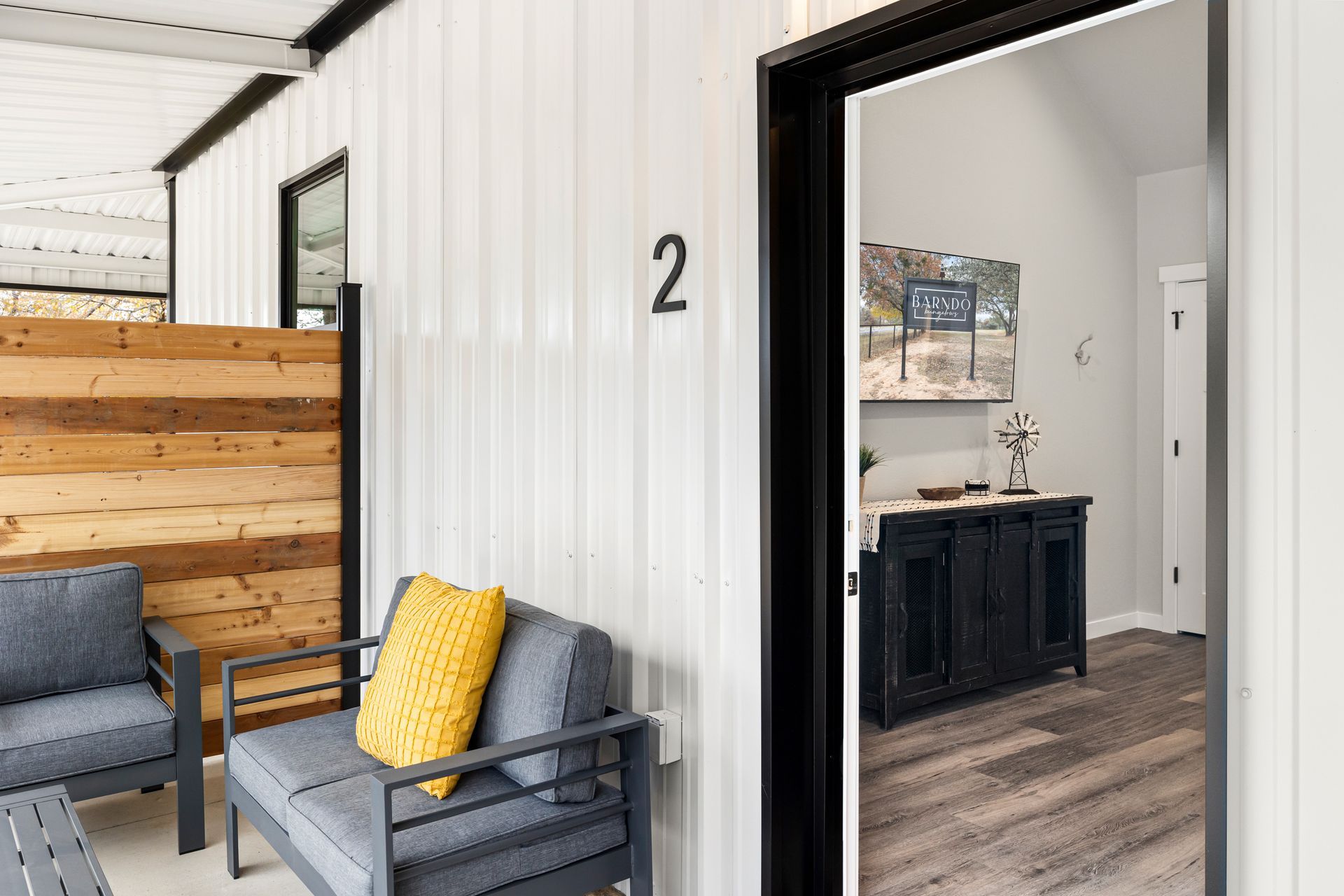 Porch seating beside a doorway marked 2, with gray chairs and a yellow pillow, looking into a room with a dark cabinet