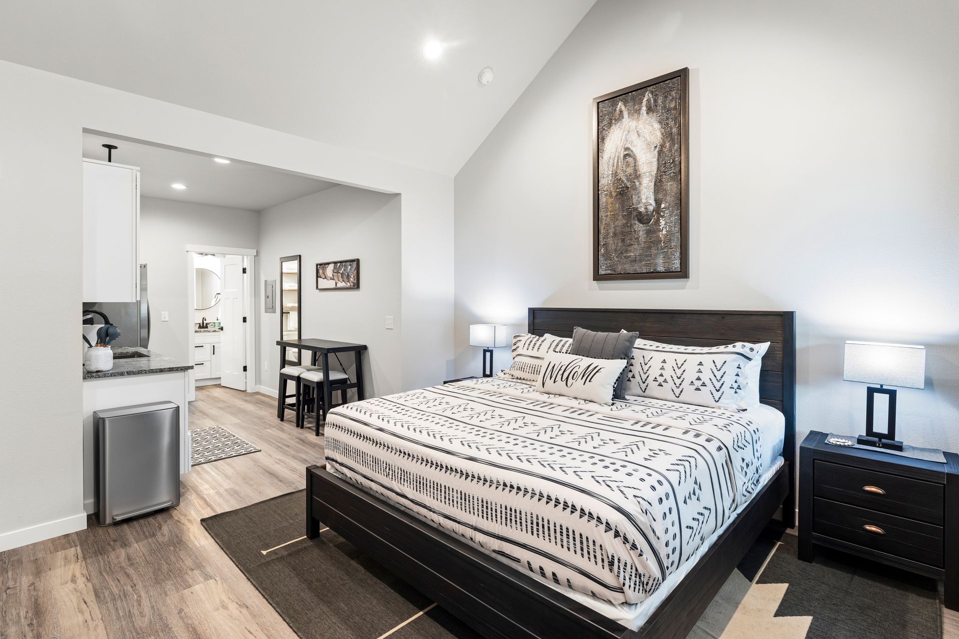 Modern bedroom with a black bed, white walls, nightstands, and adjoining kitchenette in the background