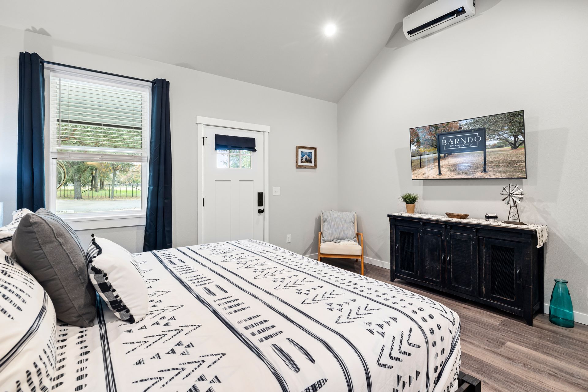 Bright bedroom with a patterned bed, white walls, window, door, and black dresser beneath wall art.