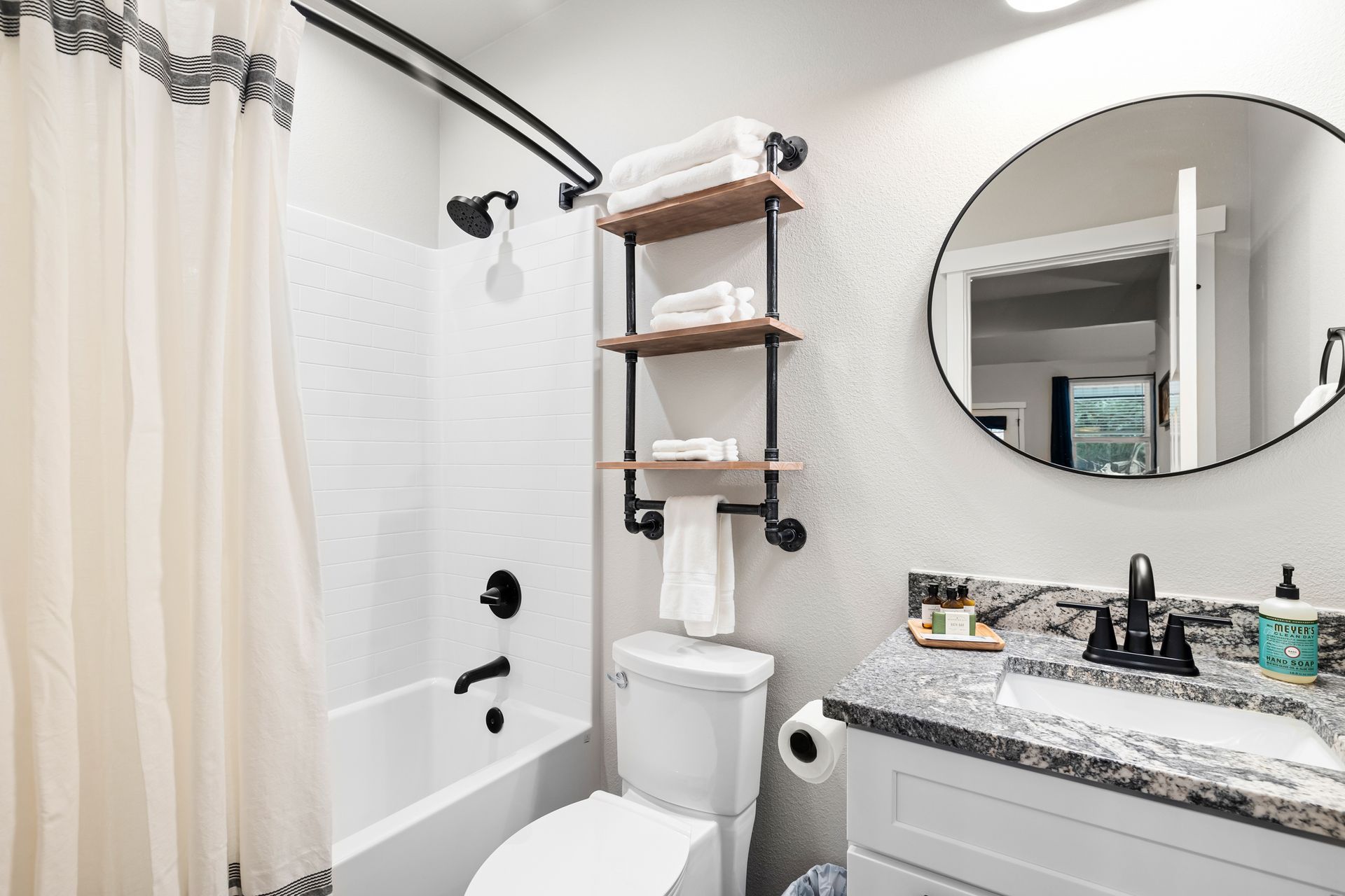 Small bathroom with shower, toilet, vanity sink, round mirror, and wall shelves.