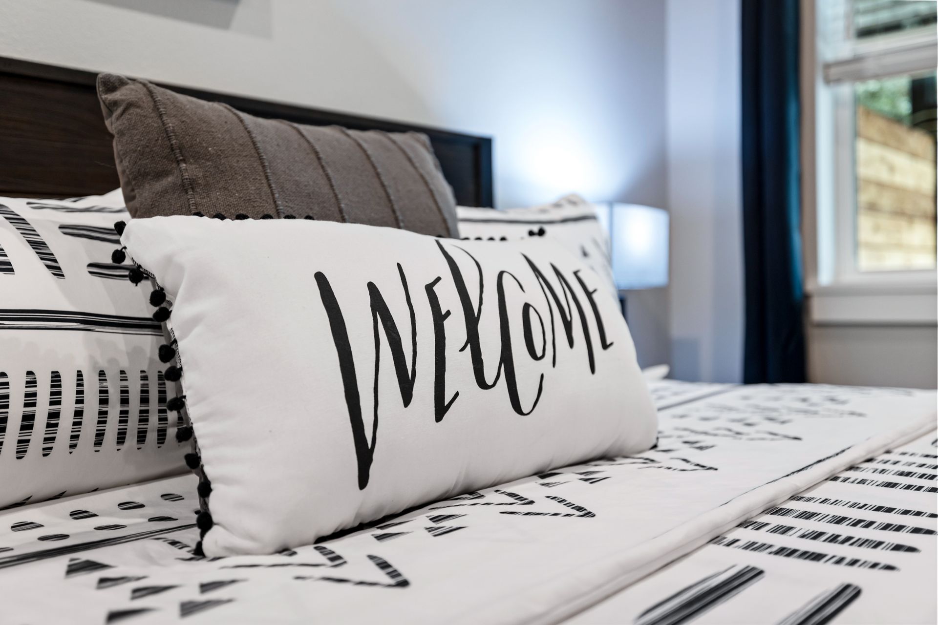Stylish bed with white and gray bedding and a decorative “Welcome” pillow in a bright bedroom.