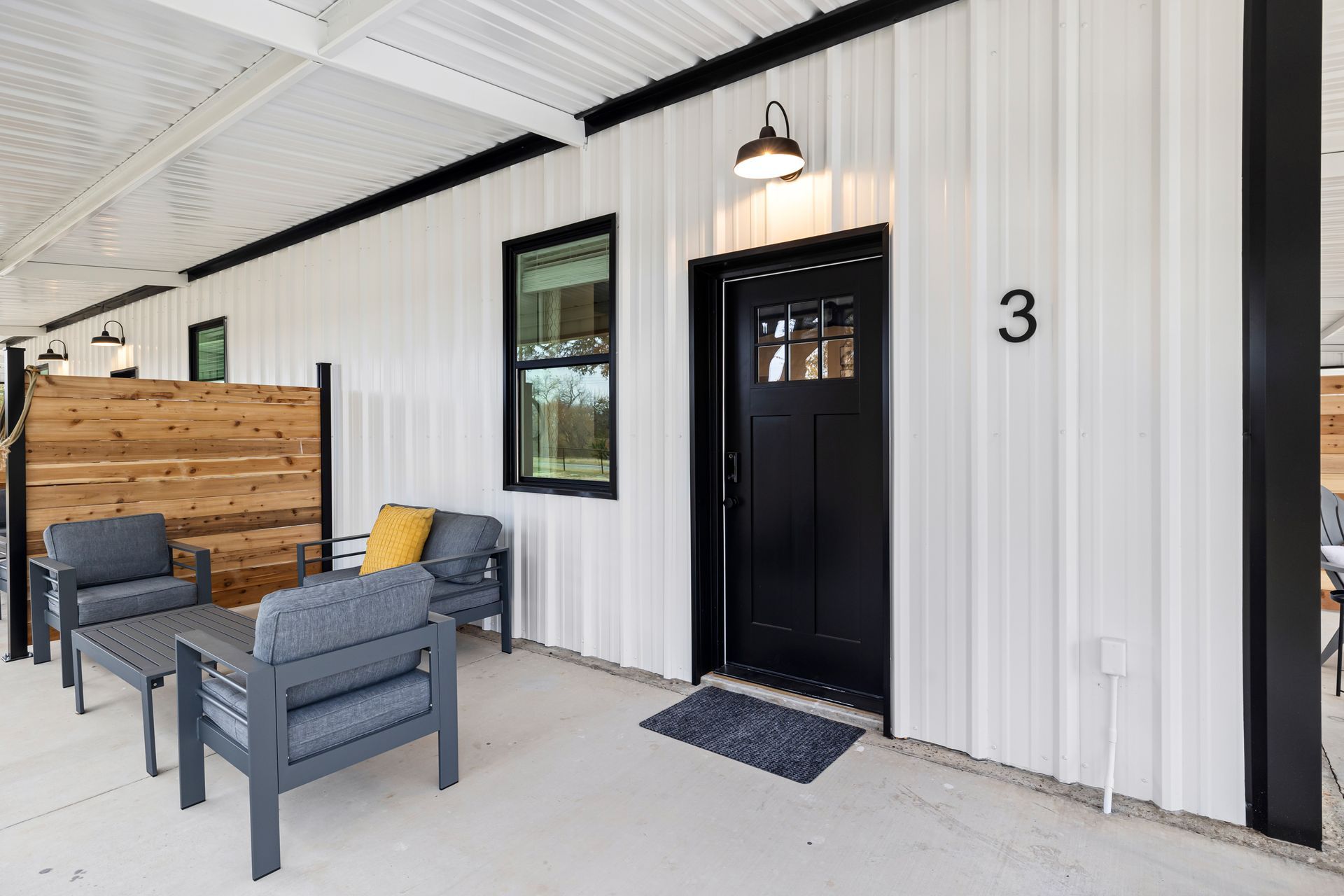 Covered porch with black door, gray chairs, wood fence, and house number 3.