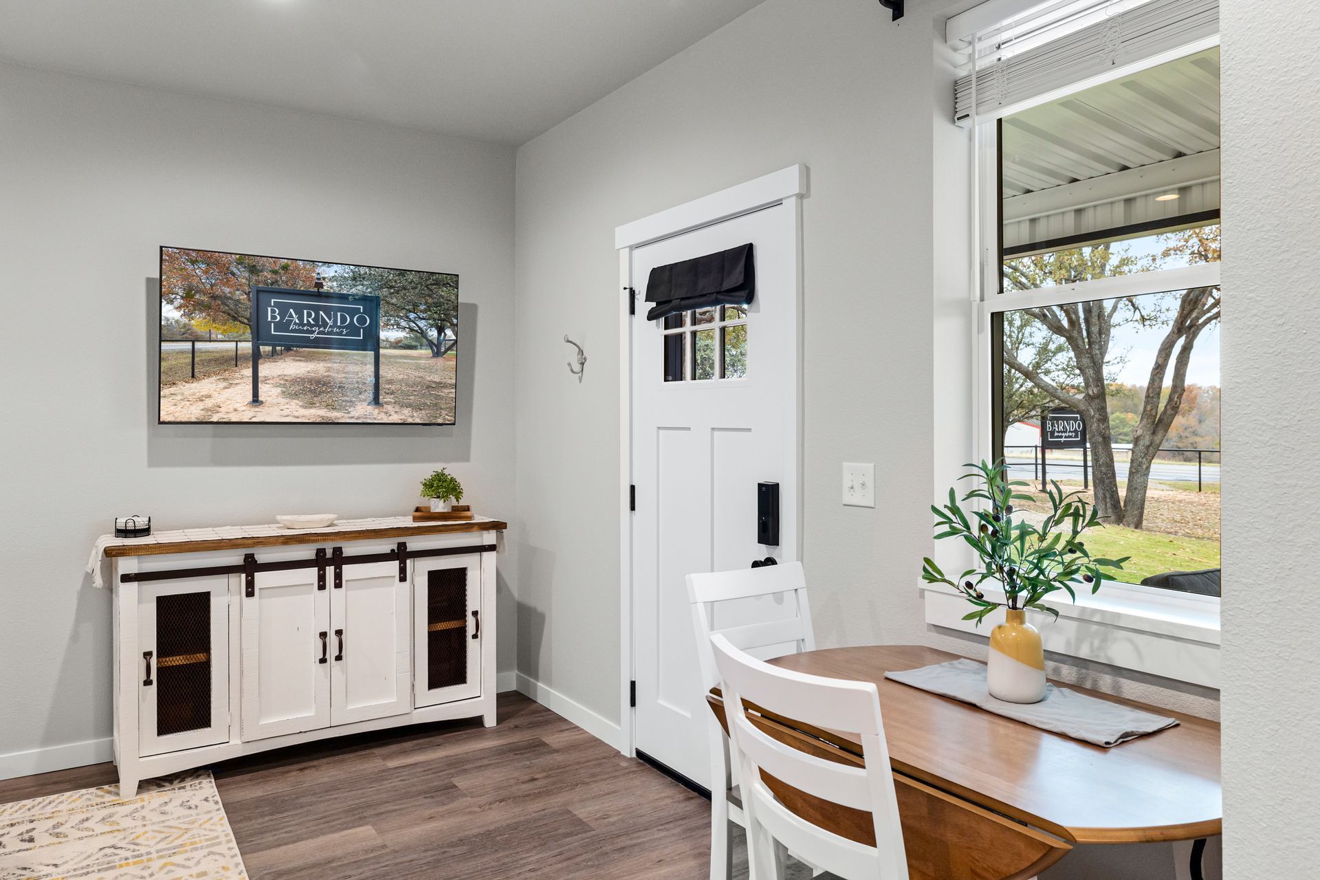 Bright entryway with white door, sideboard, wall art, and a small table by a window with a tree view