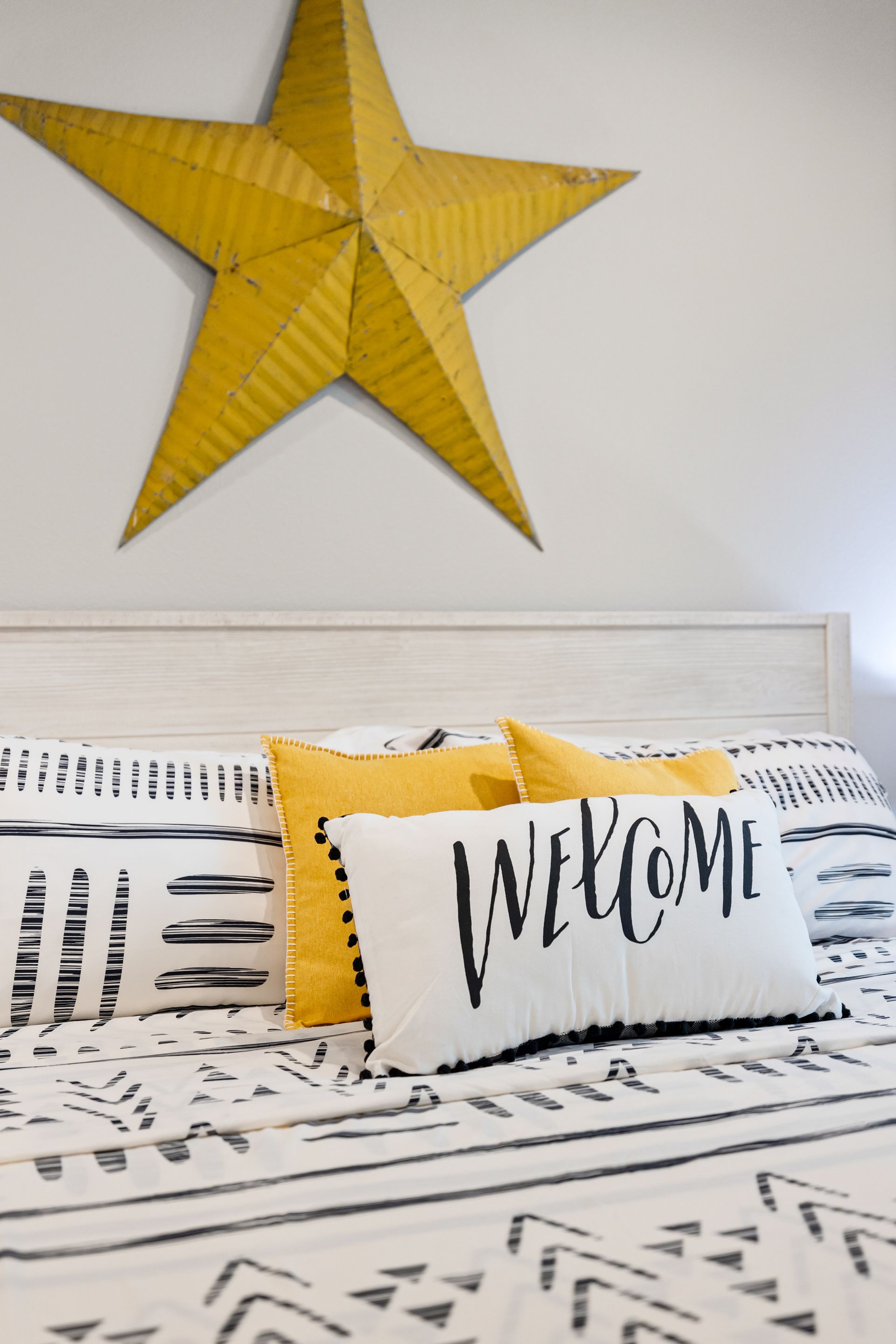 Yellow star wall decor above a bed with black-and-white bedding and a “welcome” pillow