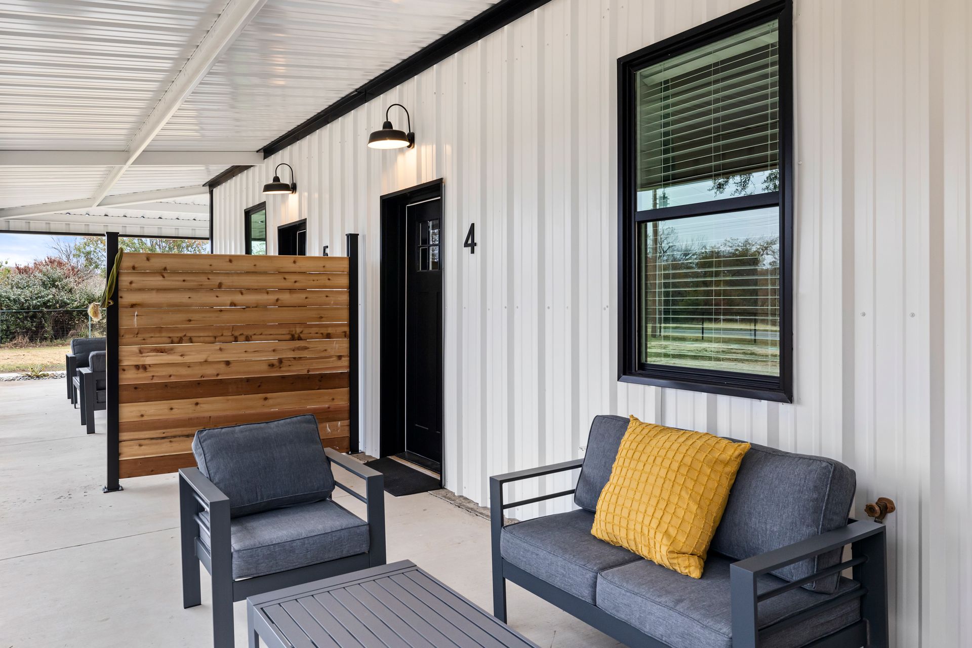 Covered porch with gray seating, yellow pillow, black door, and wood privacy screen on a white exterior wall