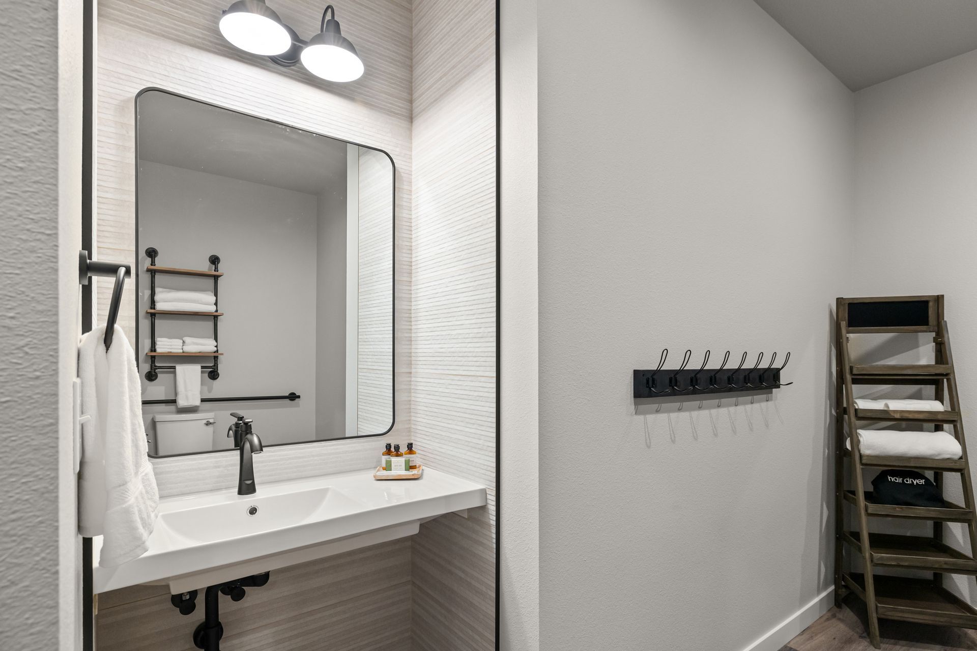 Modern bathroom with large mirror, white sink, wall lights, and a ladder-style shelf beside a tiled wall.
