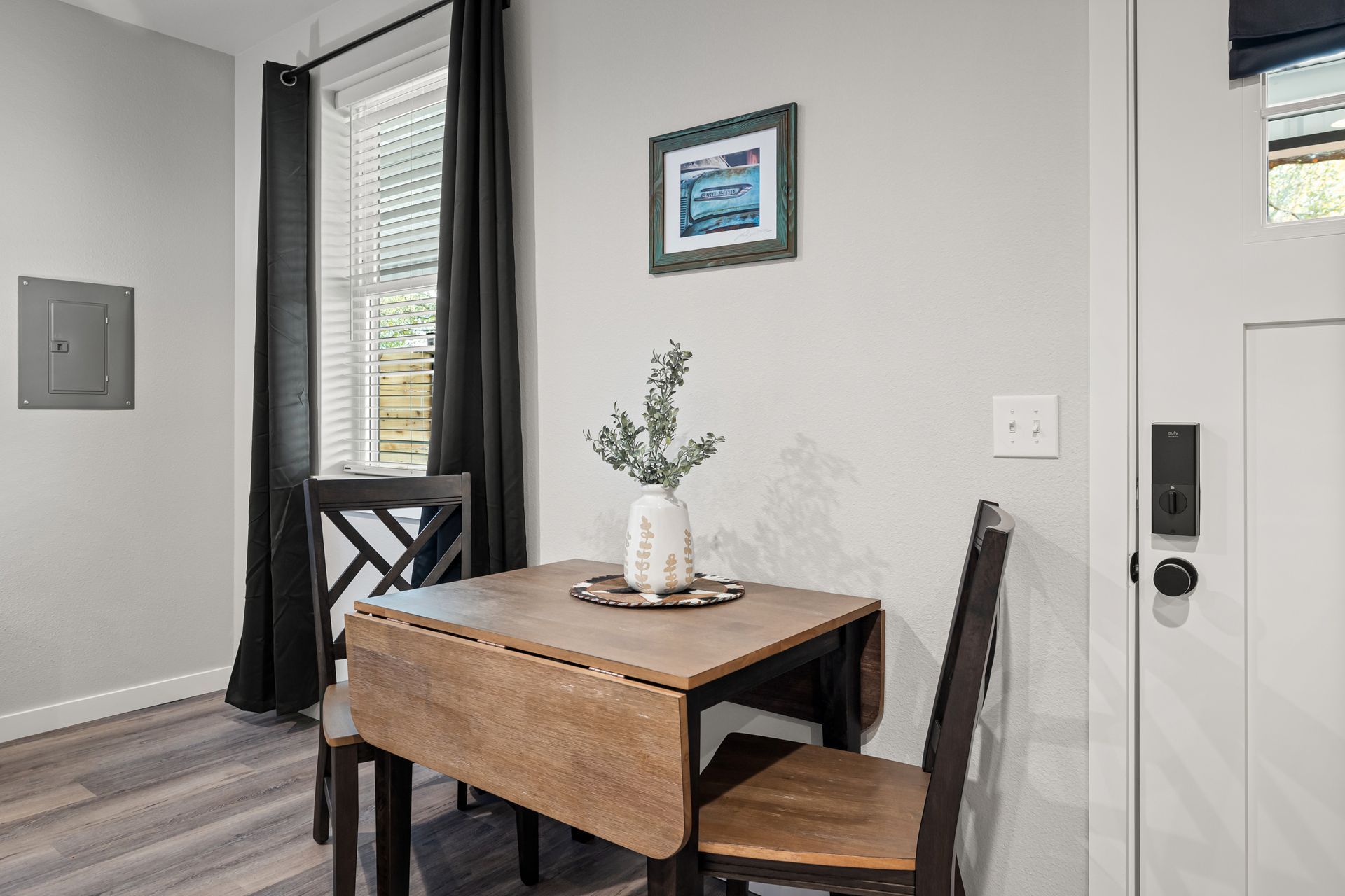 Small entryway with a wooden table, two chairs, wall art, and a black curtain by a bright window.
