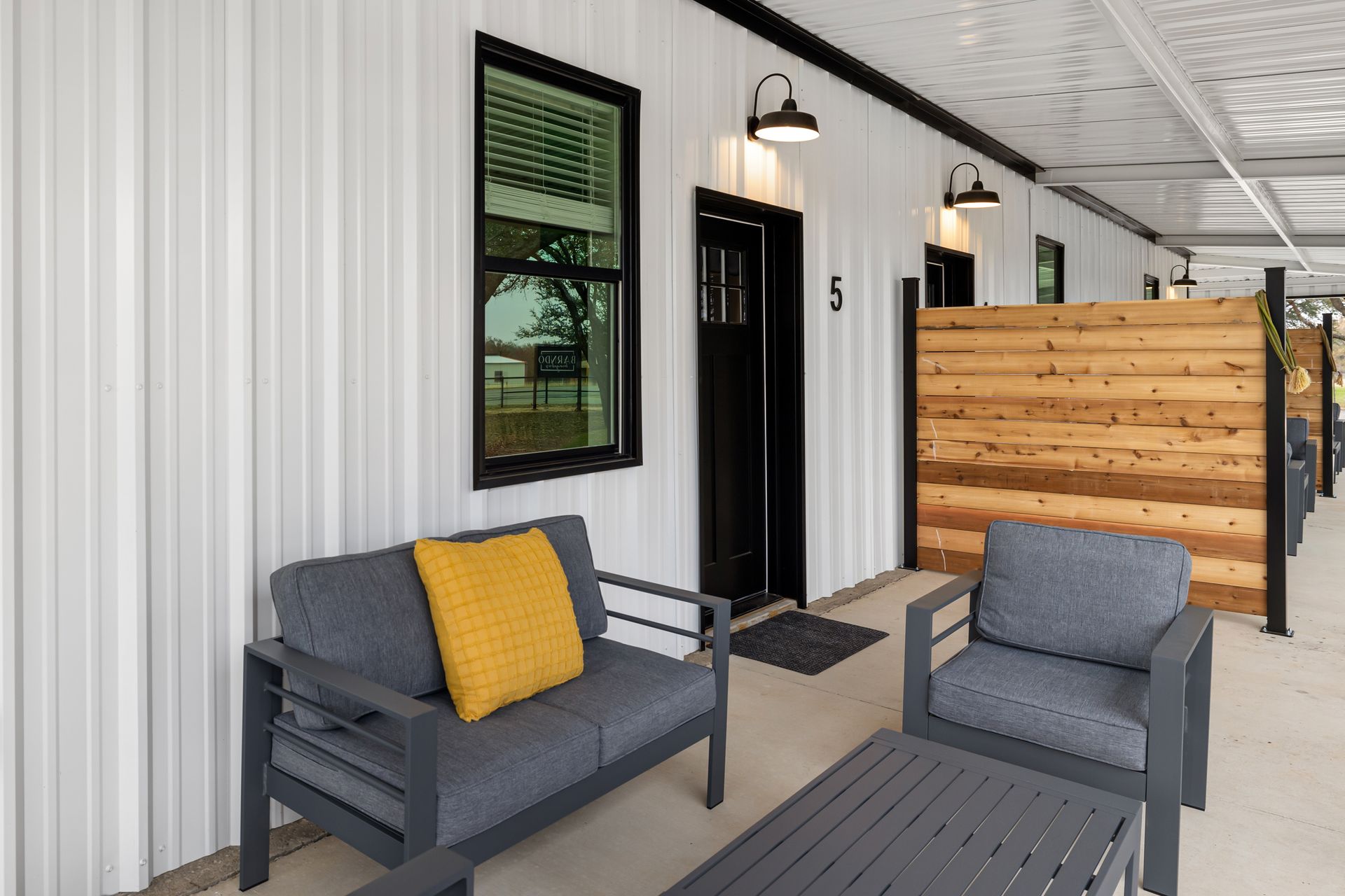 Covered front porch with gray chairs, yellow pillow, black door, and wood privacy screen