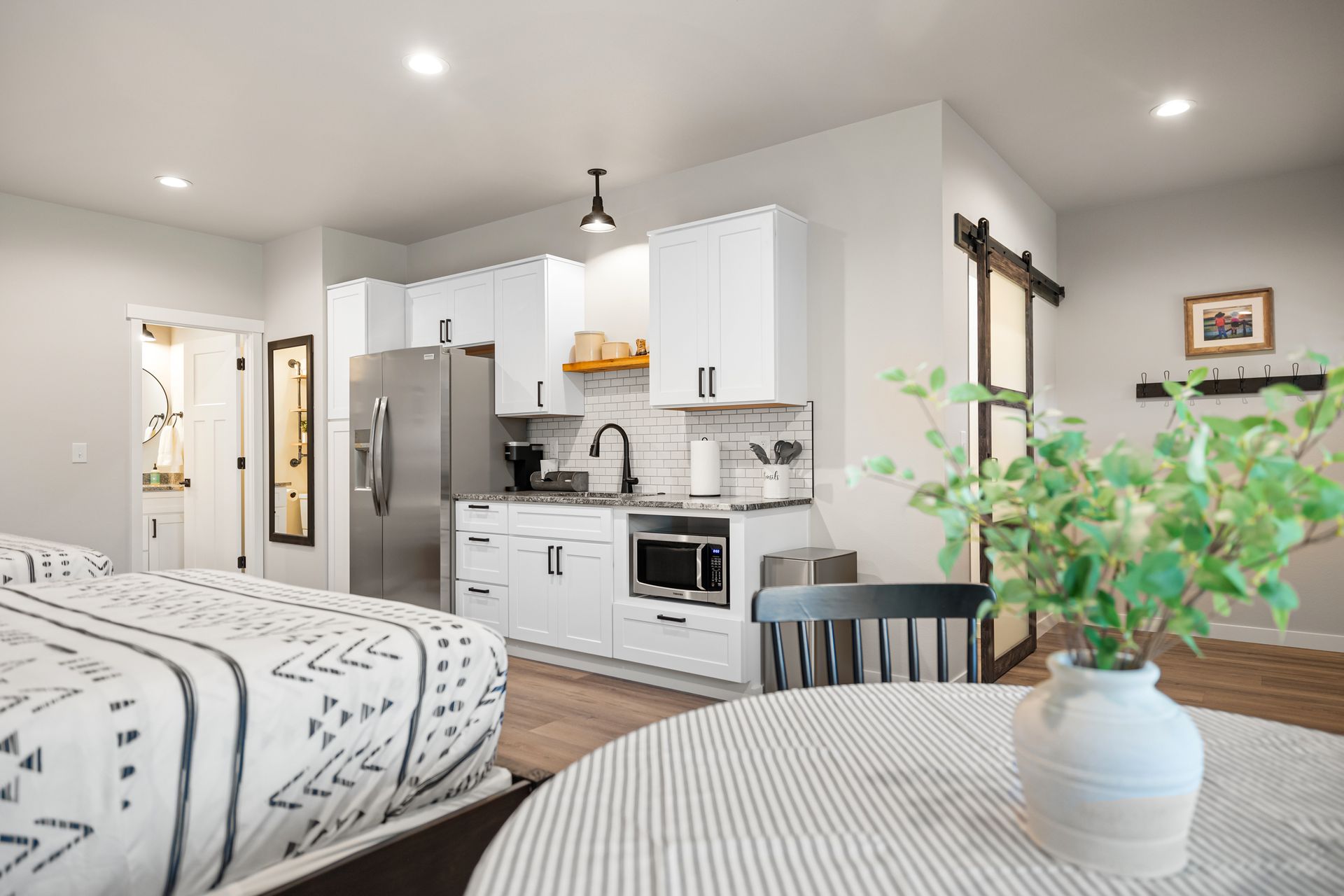 Bright studio apartment with bed, kitchen, and round table, featuring white cabinets and stainless-steel appliances