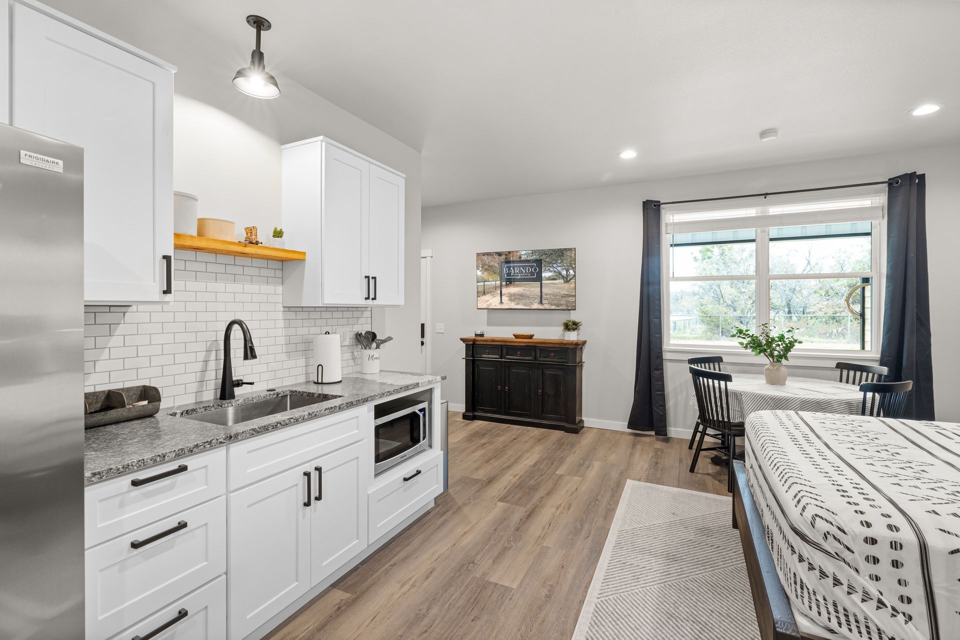 Bright studio with white kitchen, wood floor, bed, and a window with dark curtains