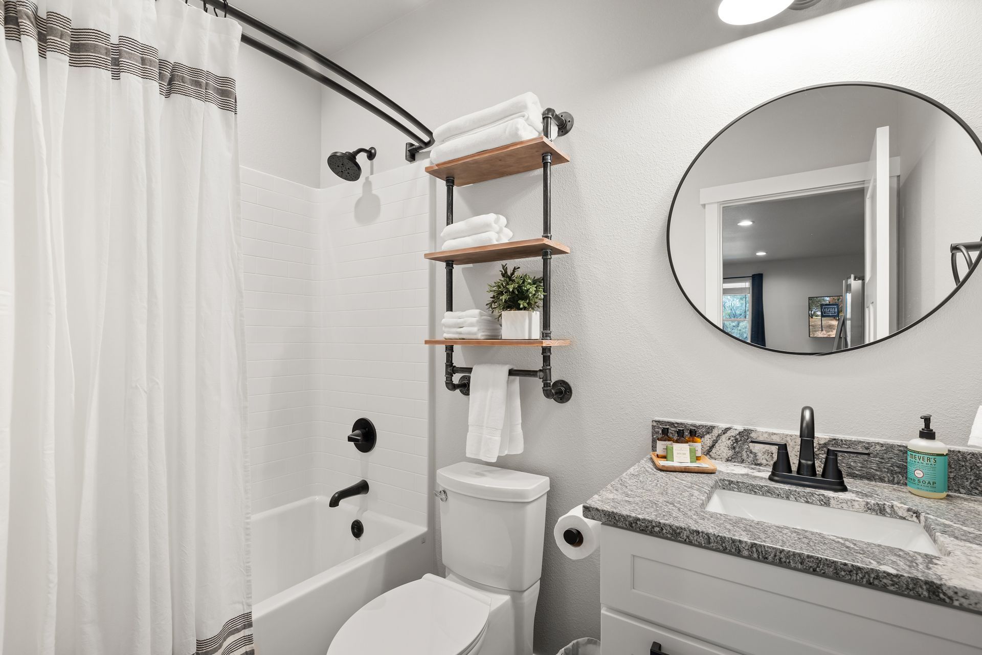 Small modern bathroom with white shower curtain, toilet, vanity, round mirror, and wall shelves